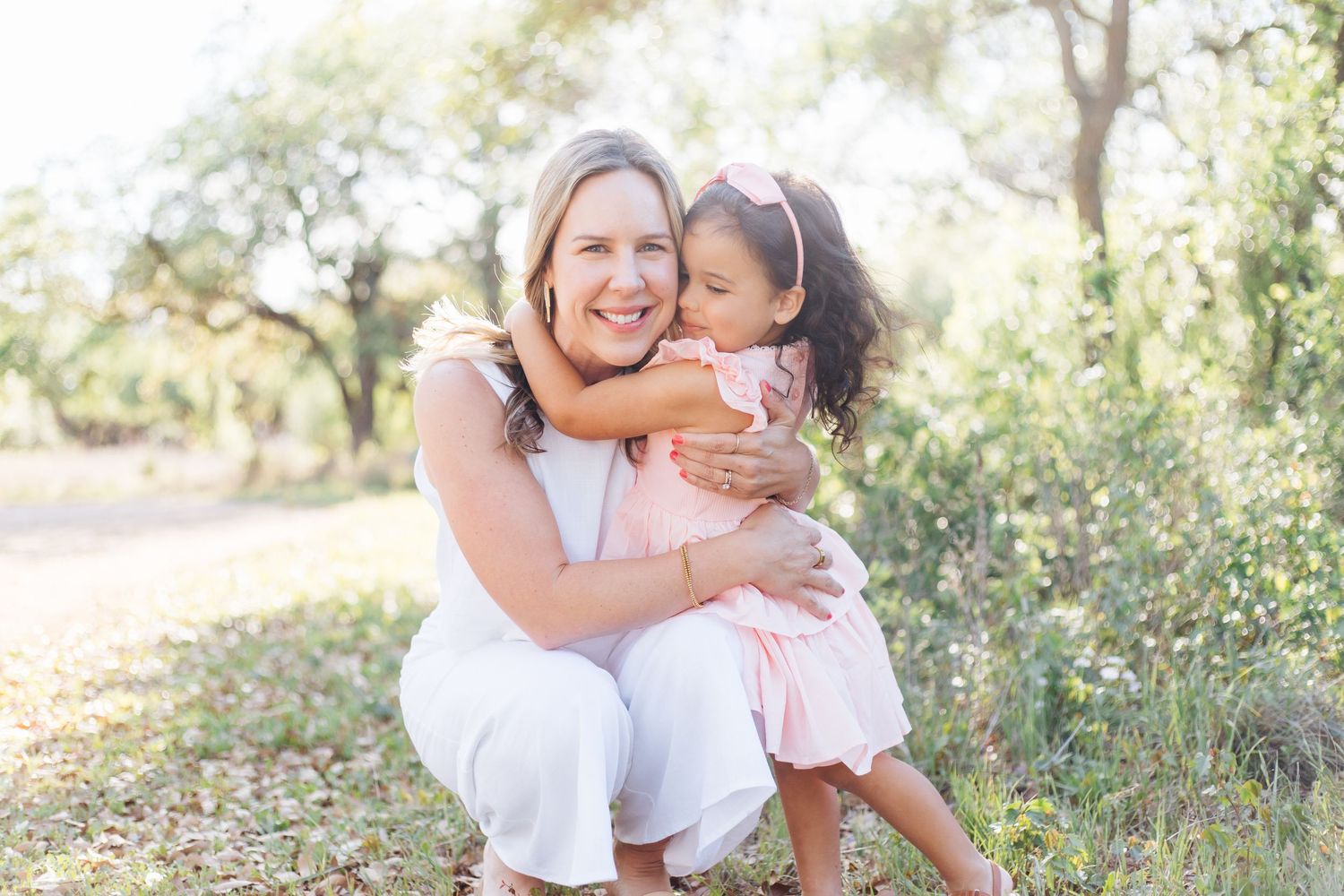 Woman in white dress hugging a young girl in pink outdoors among green trees in a bright natural setting.