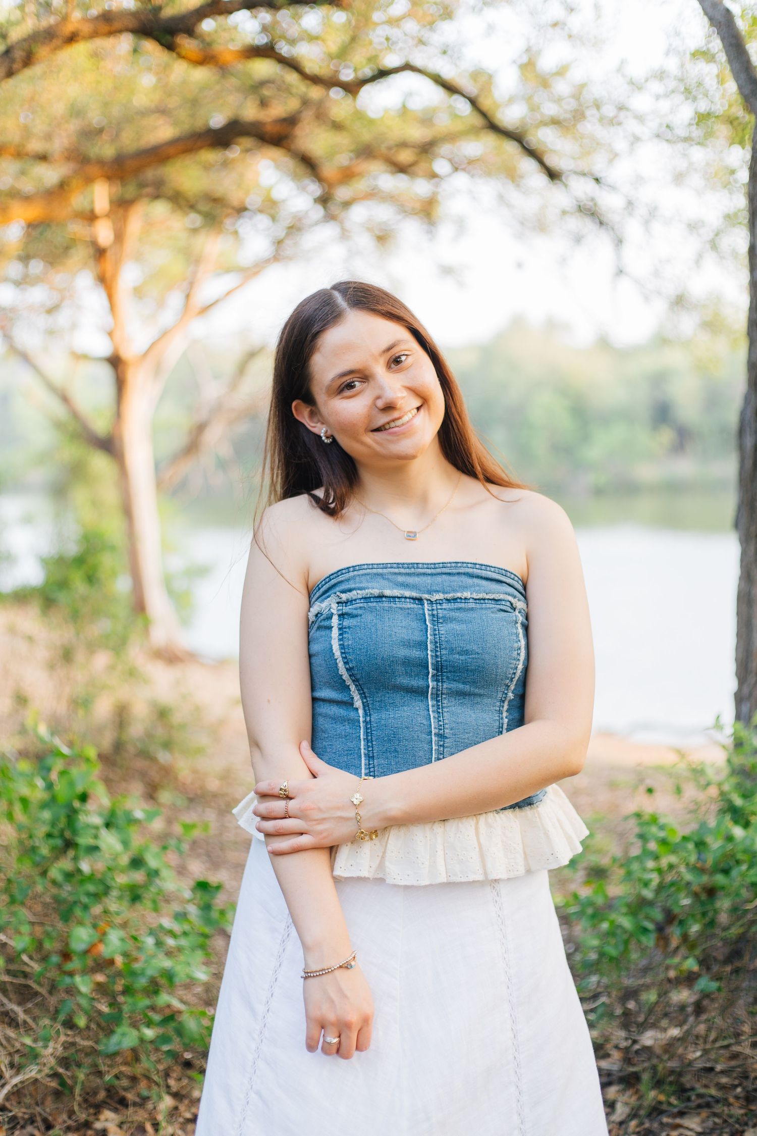 Young woman smiling in denim tube top and white skirt outdoors by a lake surrounded by trees. Senior Portraits Round Rock TX