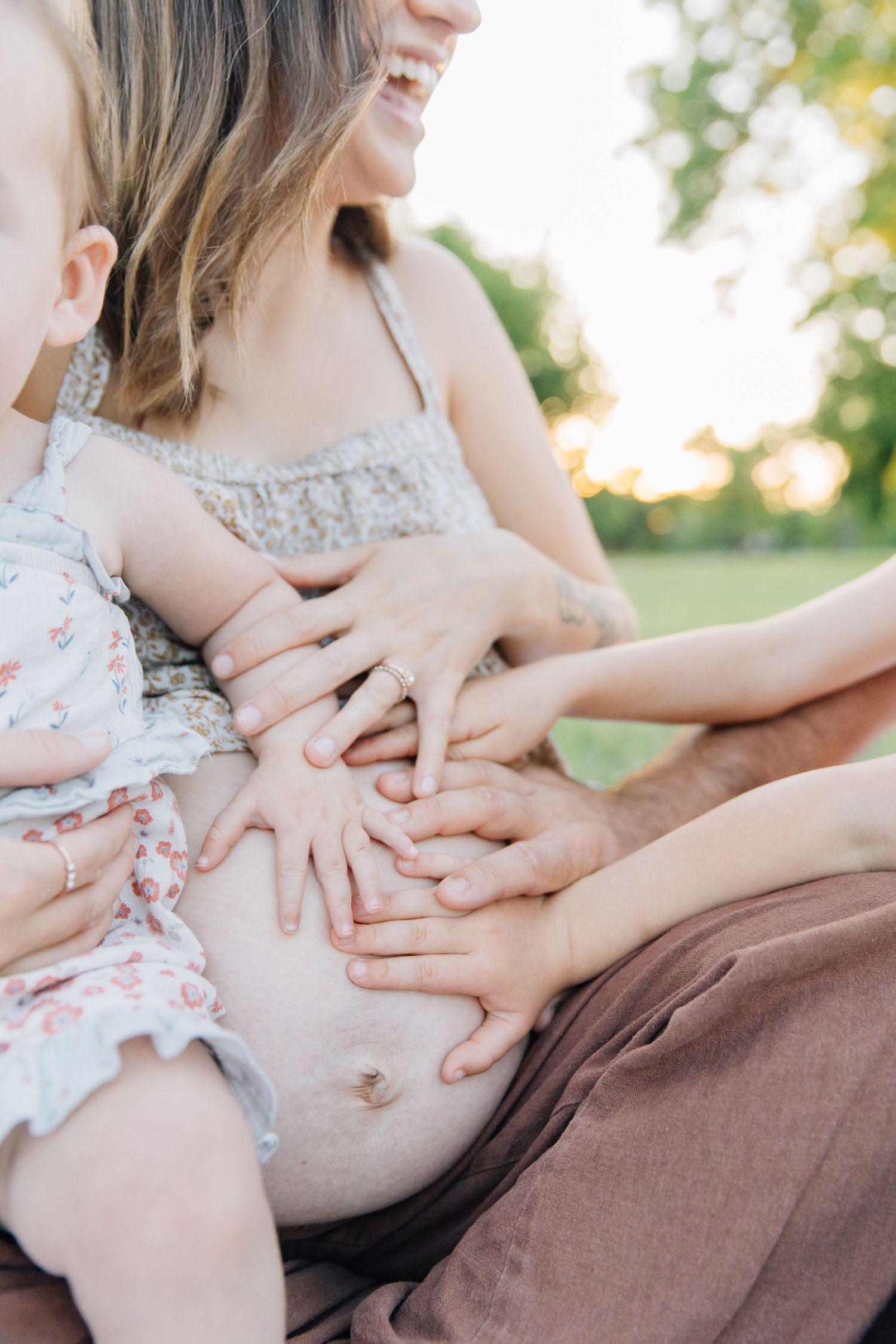 Smiling pregnant woman outdoors with children placing hands on her baby bump during a golden hour family maternity photo session.
