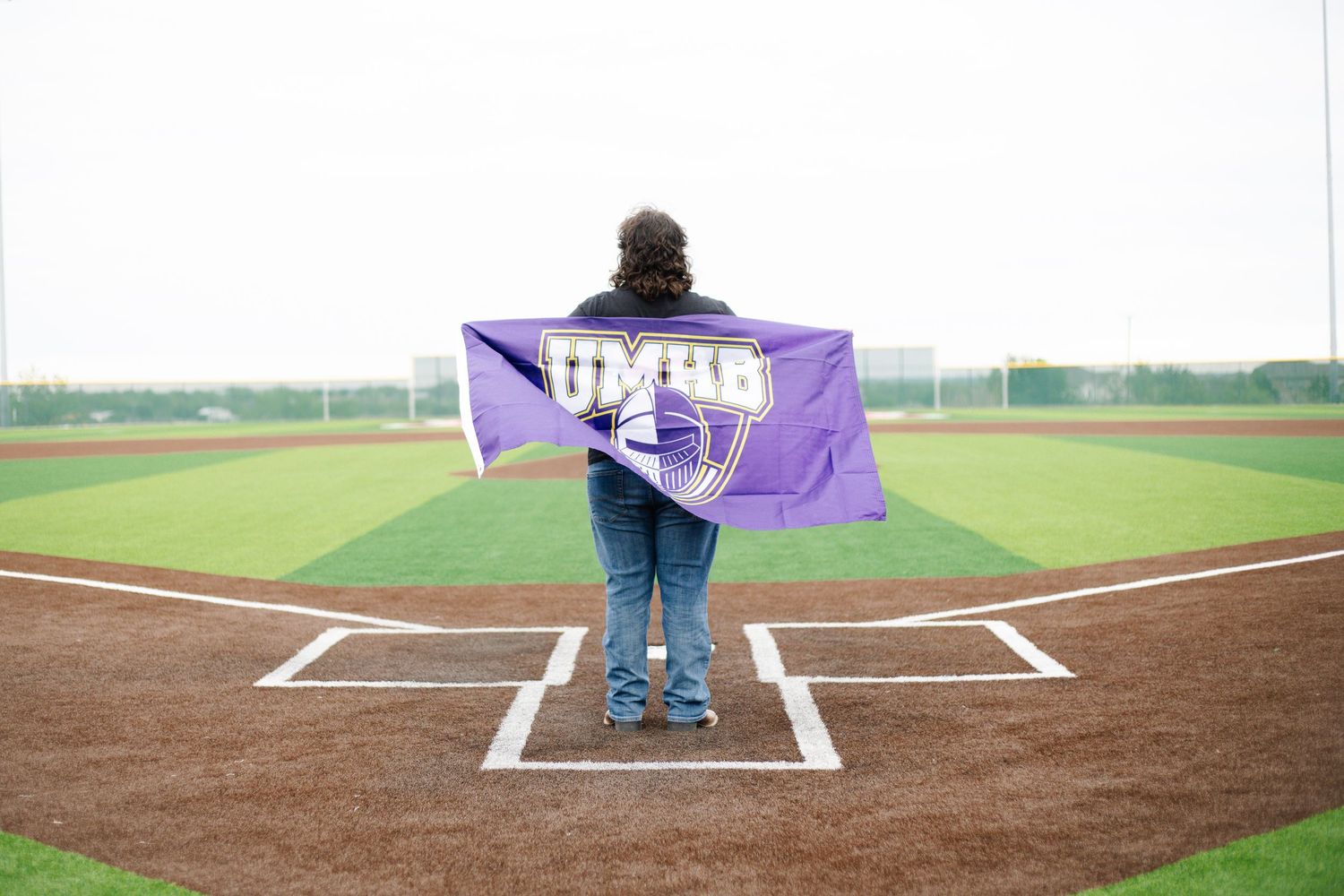 Person holding a purple UMHB flag on a baseball field home plate, with green grass and overcast sky.