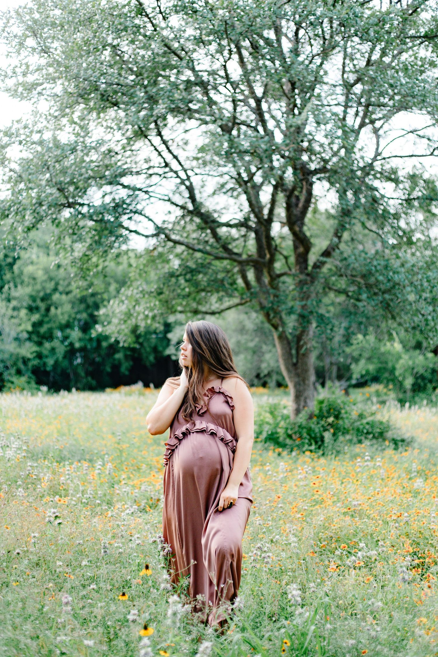 Pregnant woman in mauve dress standing in wildflower meadow with trees, cradling baby bump outdoors.