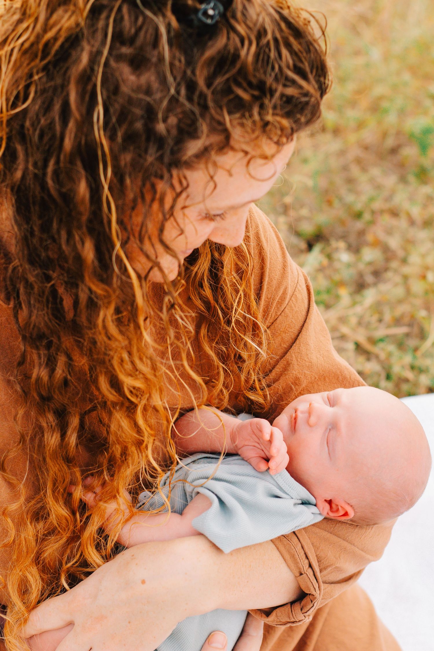Close-up of curly-haired person in brown sweater holding baby in blue outfit.