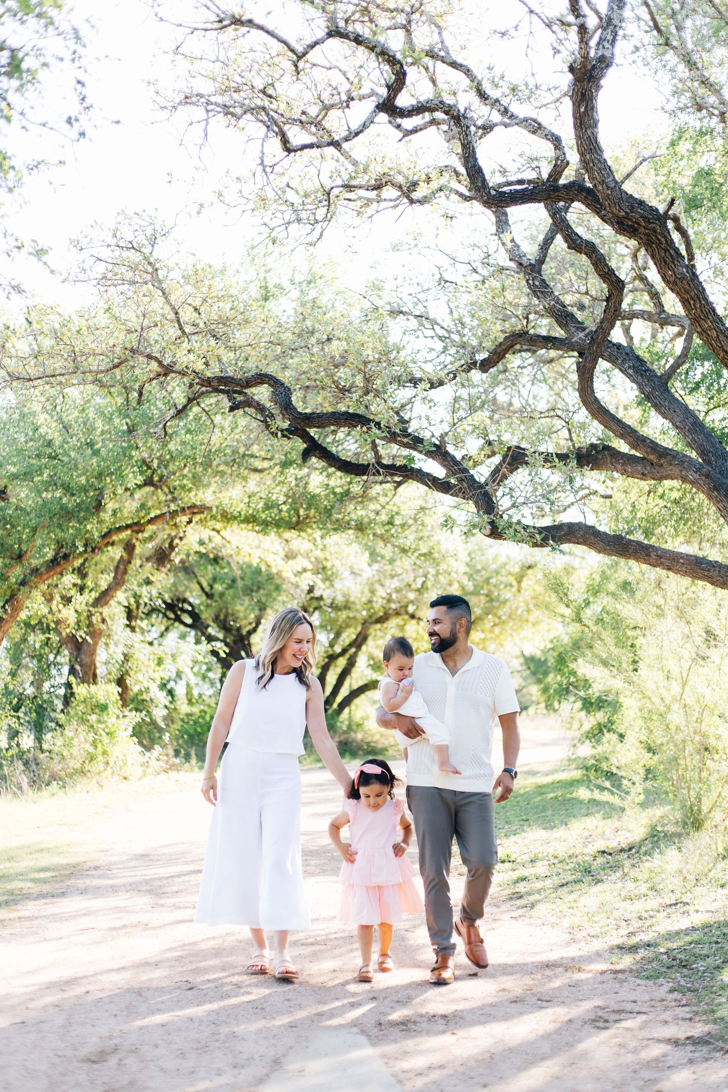 Family of four walks hand-in-hand along a sunny tree-lined dirt path in a park.