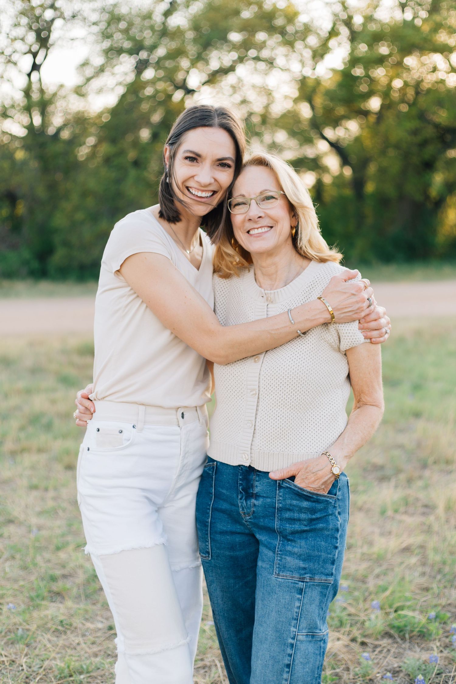 Two women smiling and hugging outdoors in a green field at golden hour.