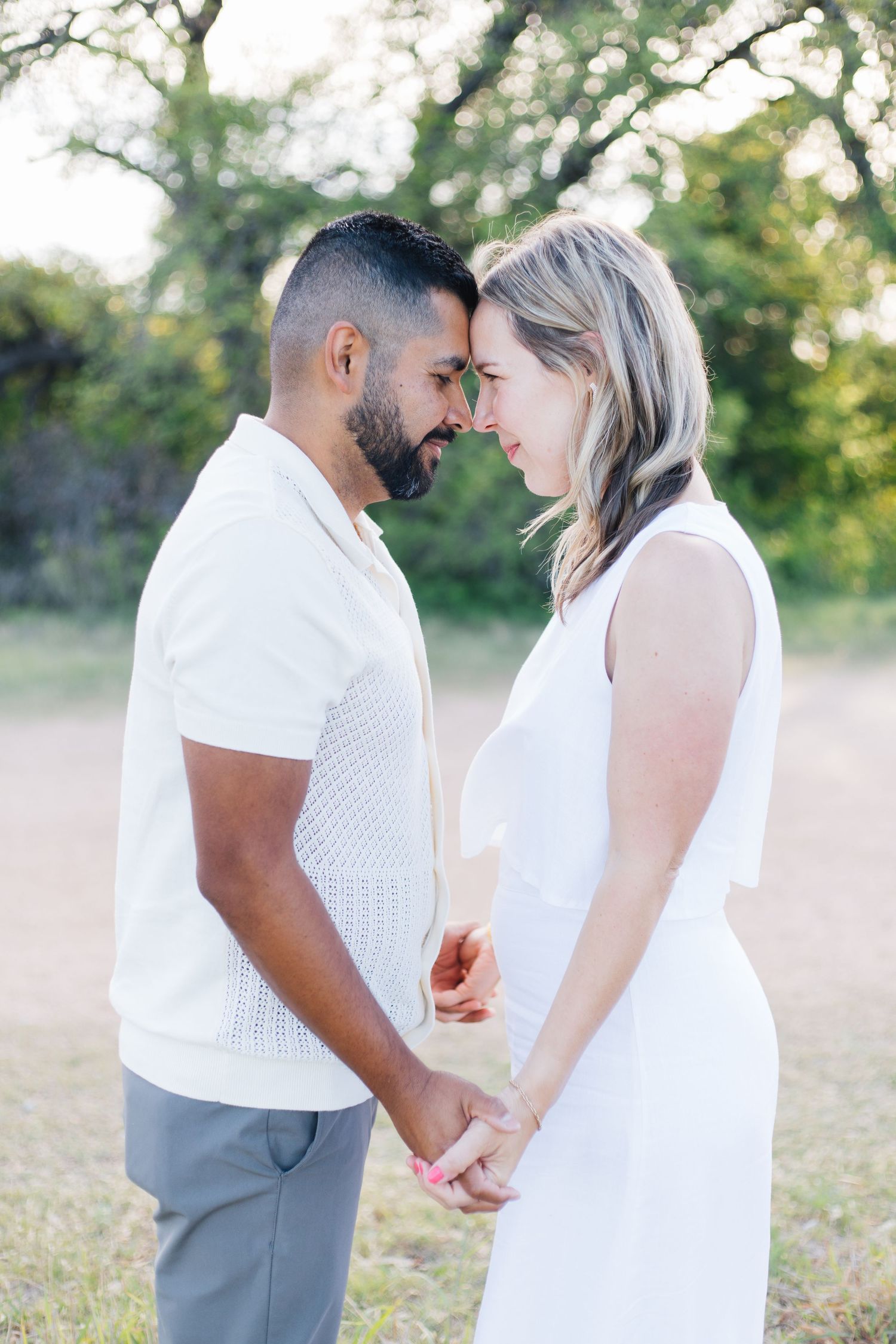 Romantic couple in white outfits holding hands and touching foreheads tenderly in a sunlit outdoor setting.