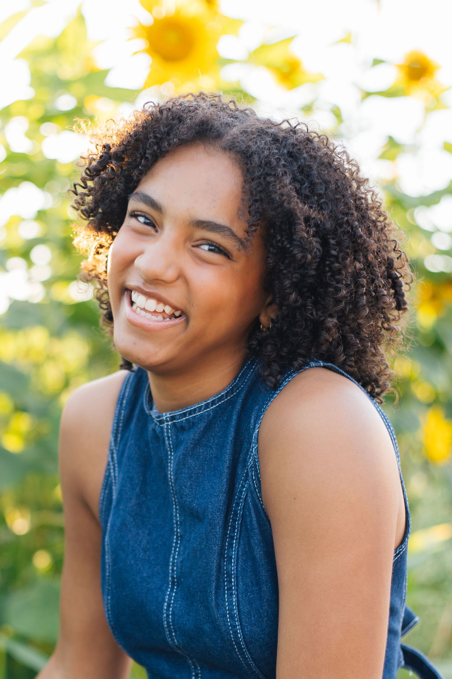 Smiling young woman with curly hair in denim top, posing outdoors in a sunflower field on a sunny day.