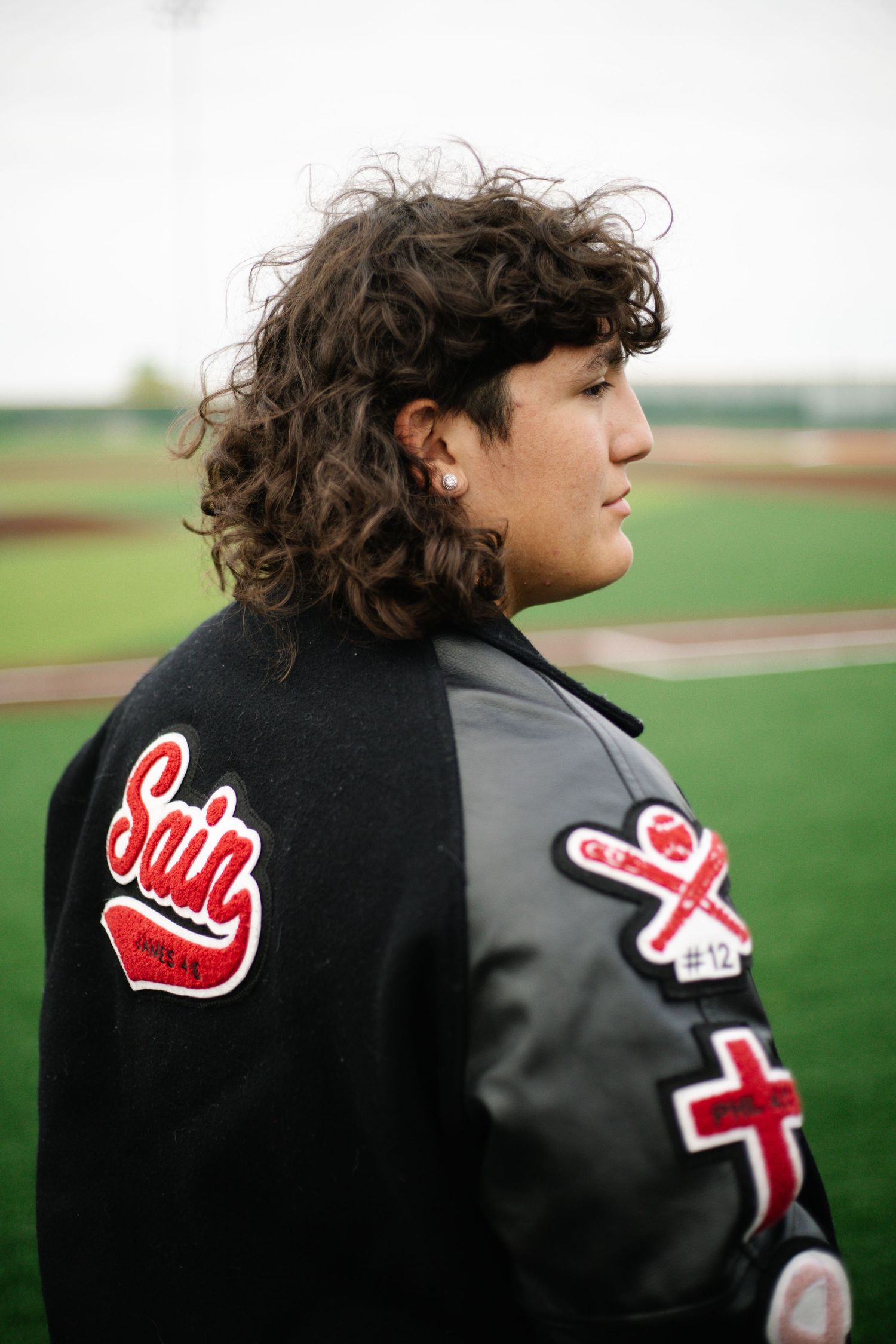 Student wearing Saints varsity letterman jacket stands on baseball field showing patches and curly hair.