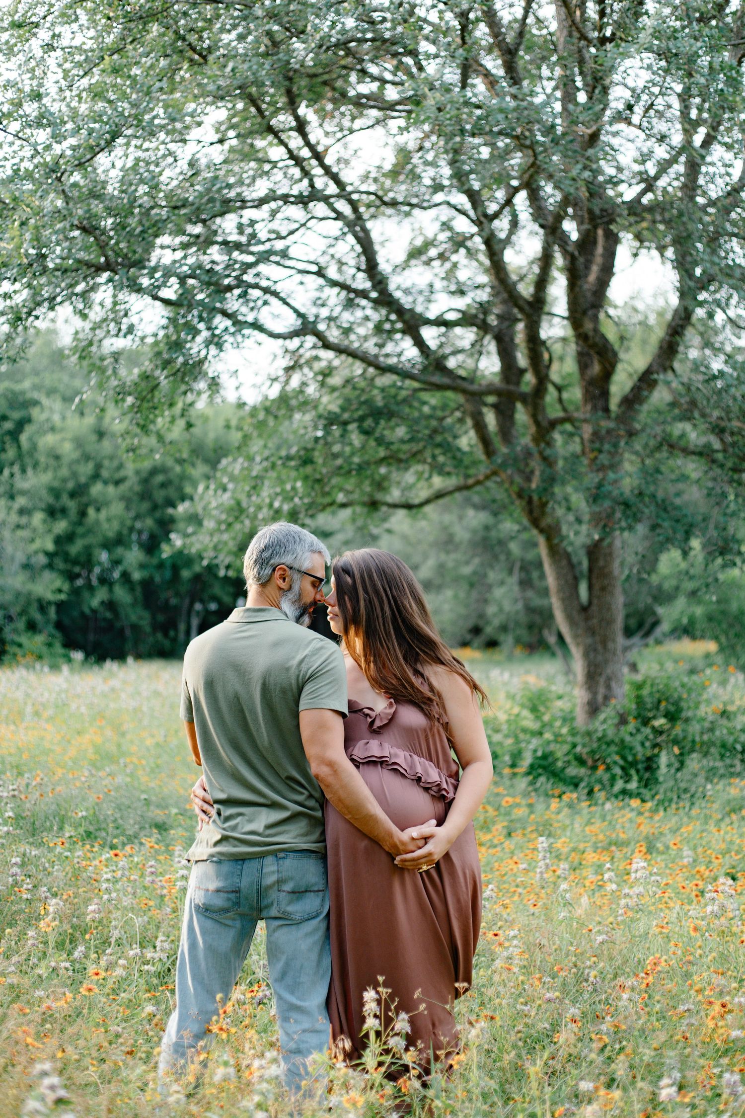 Couple embracing in a wildflower field during maternity photo session, man in green shirt with pregnant woman in brown dress.