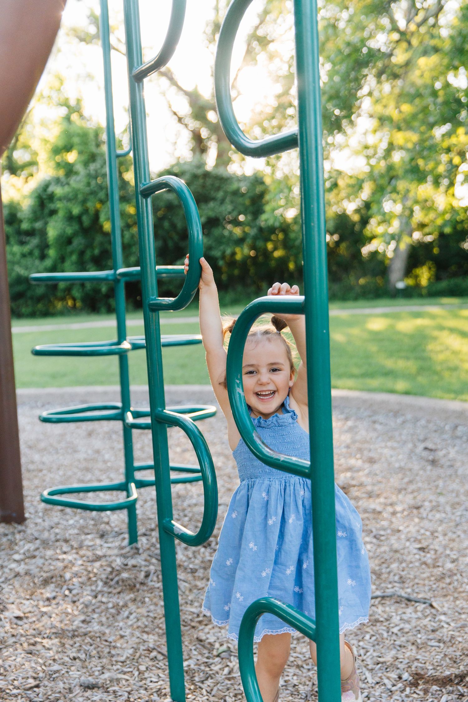 A smiling little girl in a blue dress plays on green playground monkey bars at a sunny park.