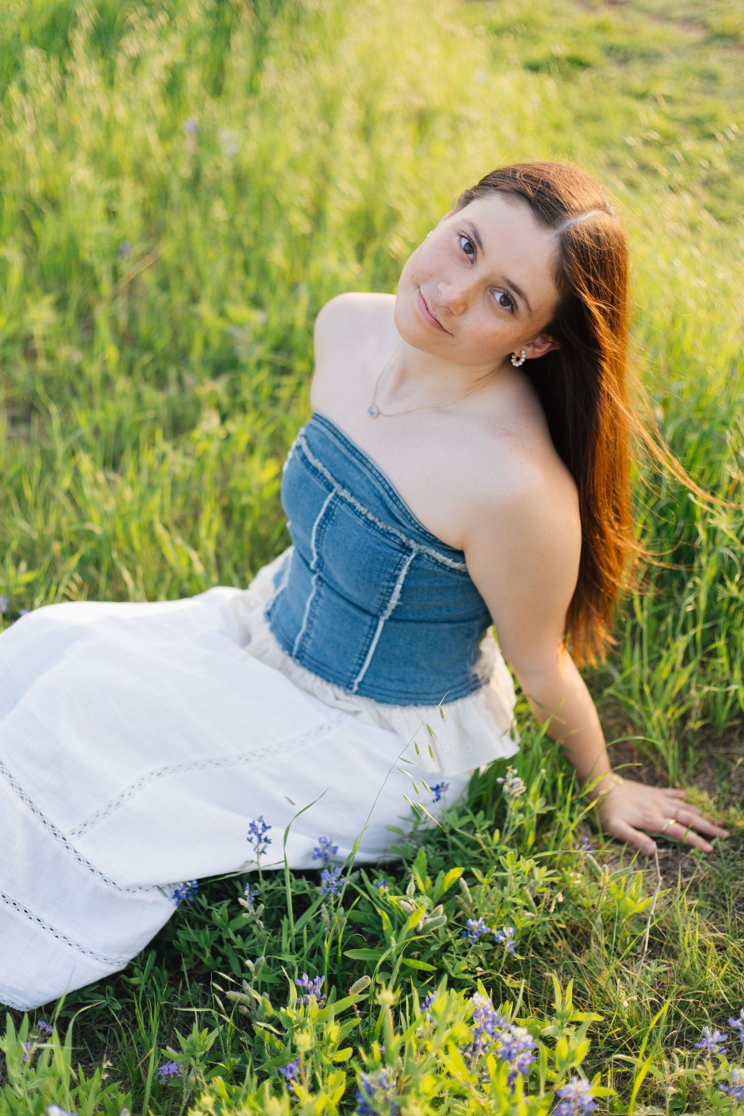 Young woman in denim corset top and white skirt sitting in green meadow with wildflowers, smiling outdoors. Senior Portraits Round Rock TX