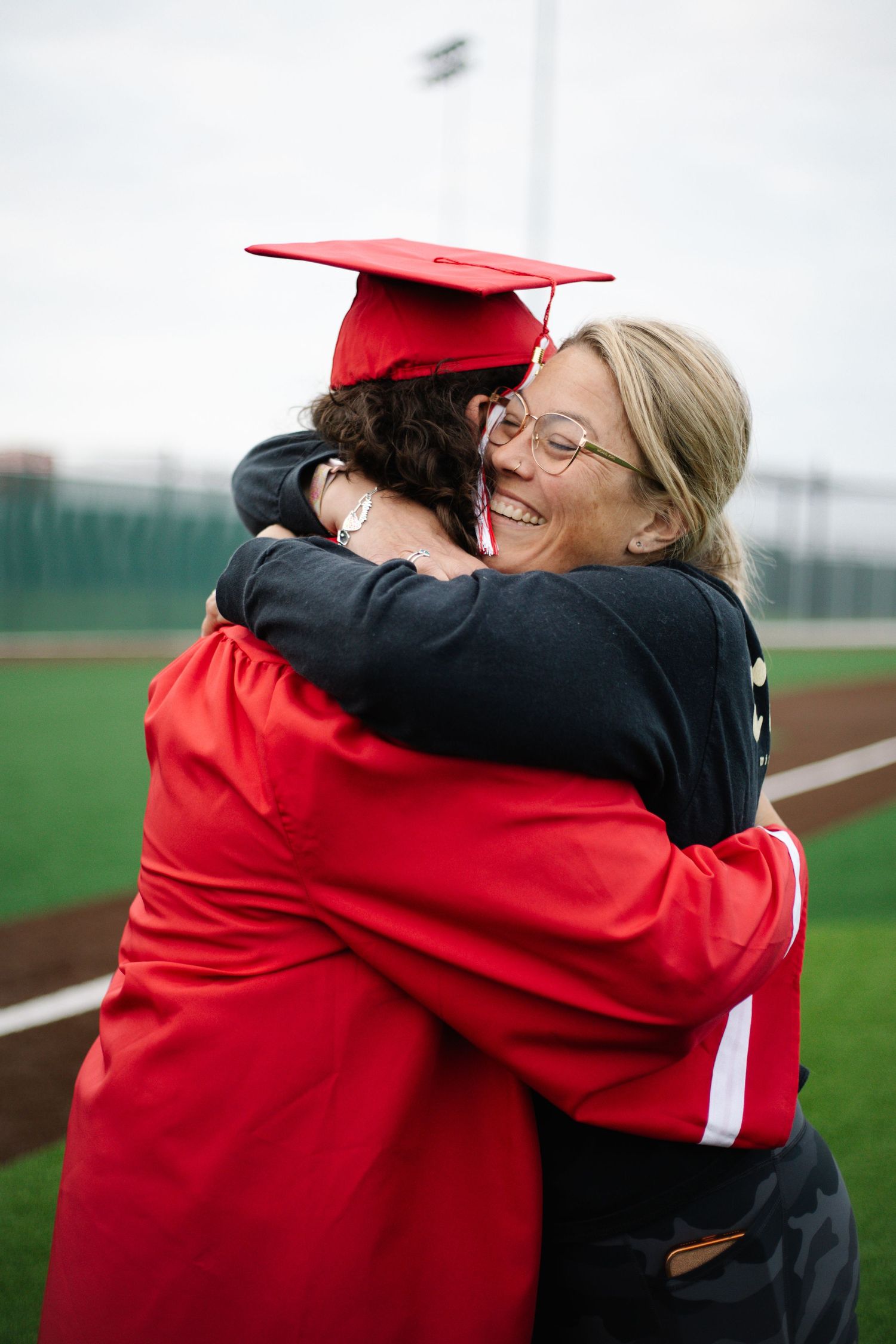 Graduate in red cap and gown shares joyful hug with smiling blonde woman on baseball field.