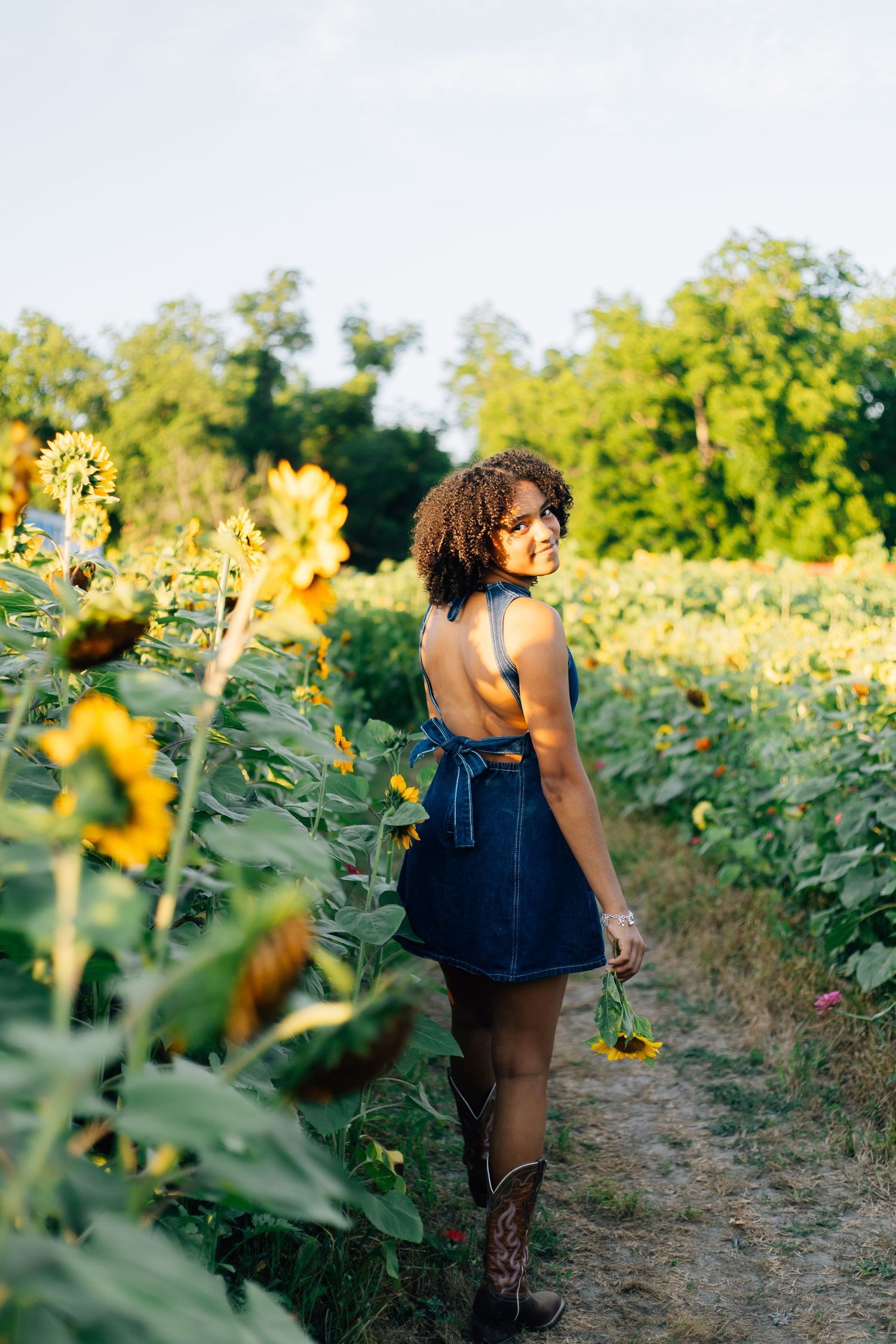 Young woman in a backless denim dress walking through a sunflower field, glancing back with a smile, wearing cowboy boots.