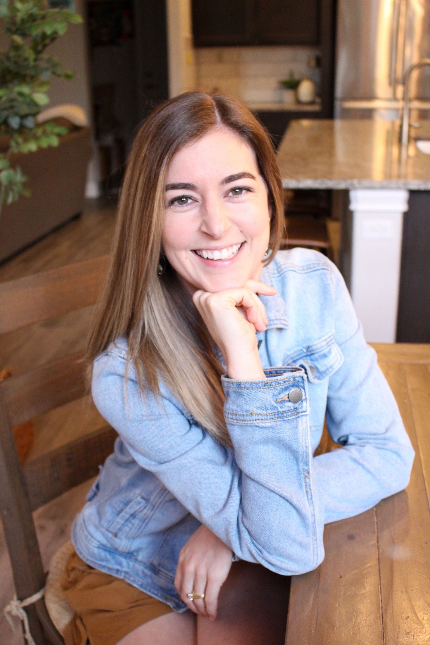 Woman in denim jacket smiles warmly while resting chin on hand in casual indoor setting.