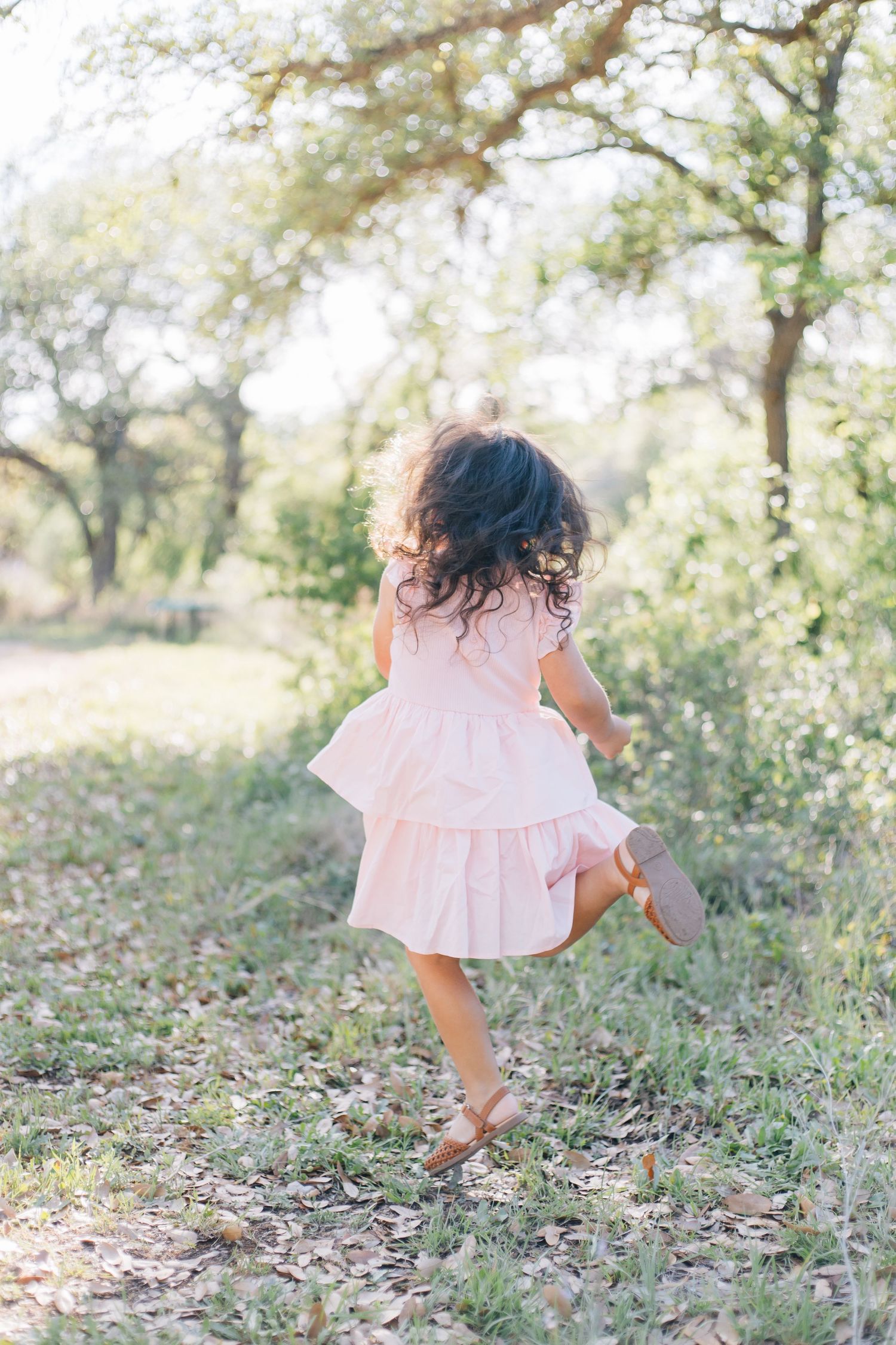 Young girl in pink ruffled dress runs through a sunlit wooded path, viewed from behind.