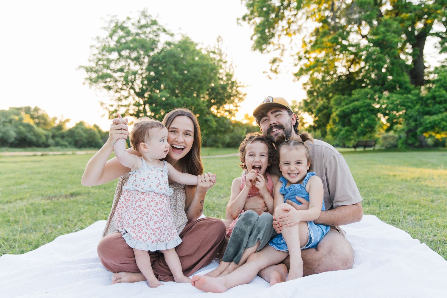 Happy family of five sitting on a white blanket in a sunny park, parents smiling with three young children.