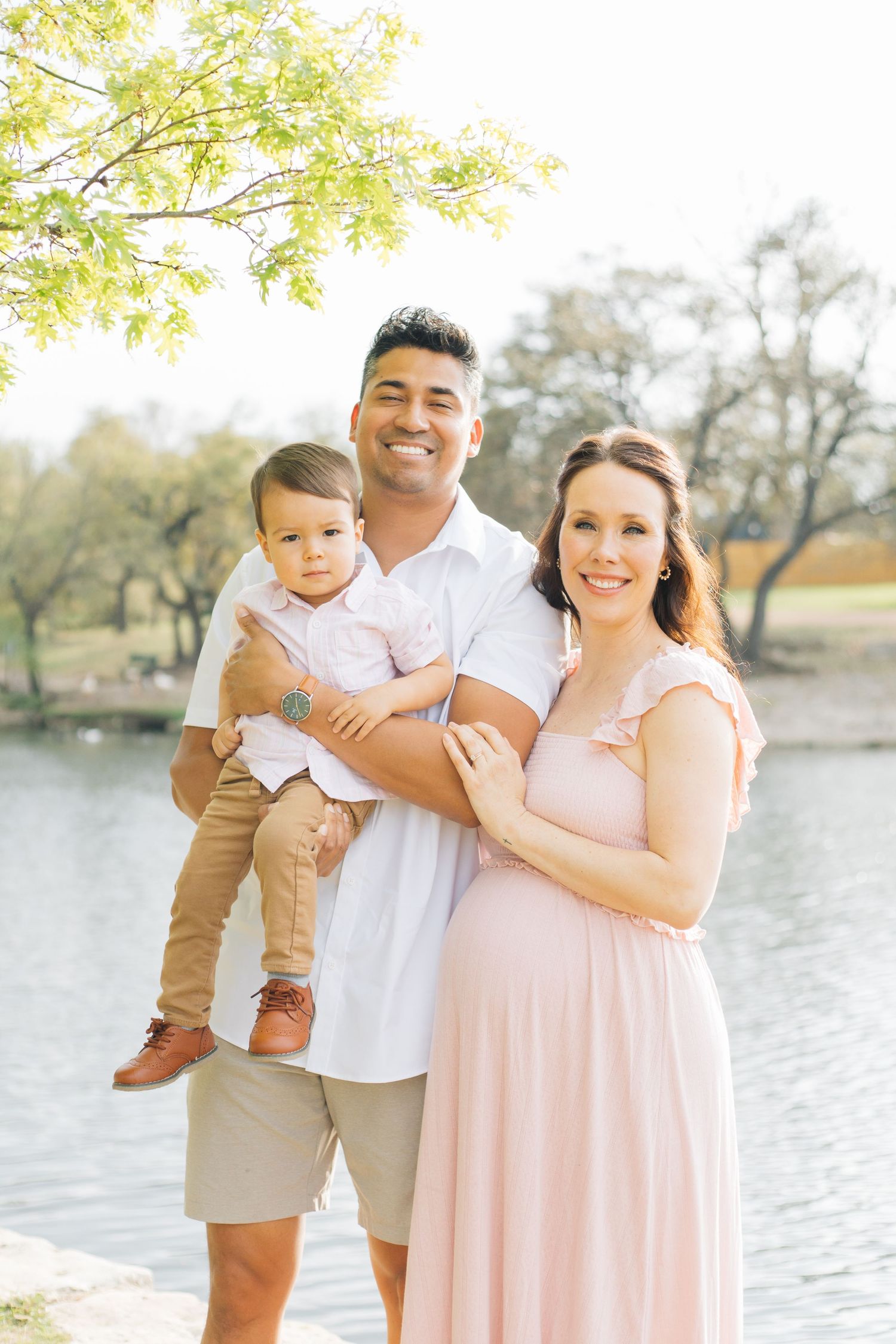 A family poses together outdoors by a lake wearing coordinating pink and white outfits on a sunny spring day.