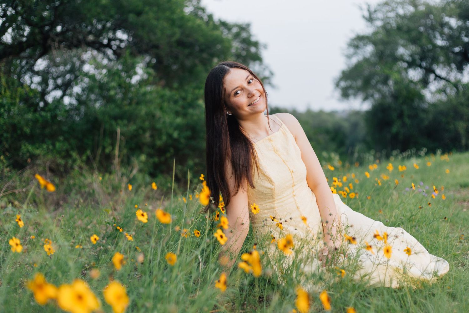 Young woman in a yellow dress sitting in a field of wildflowers, smiling softly surrounded by lush green trees. Senior Portraits TX