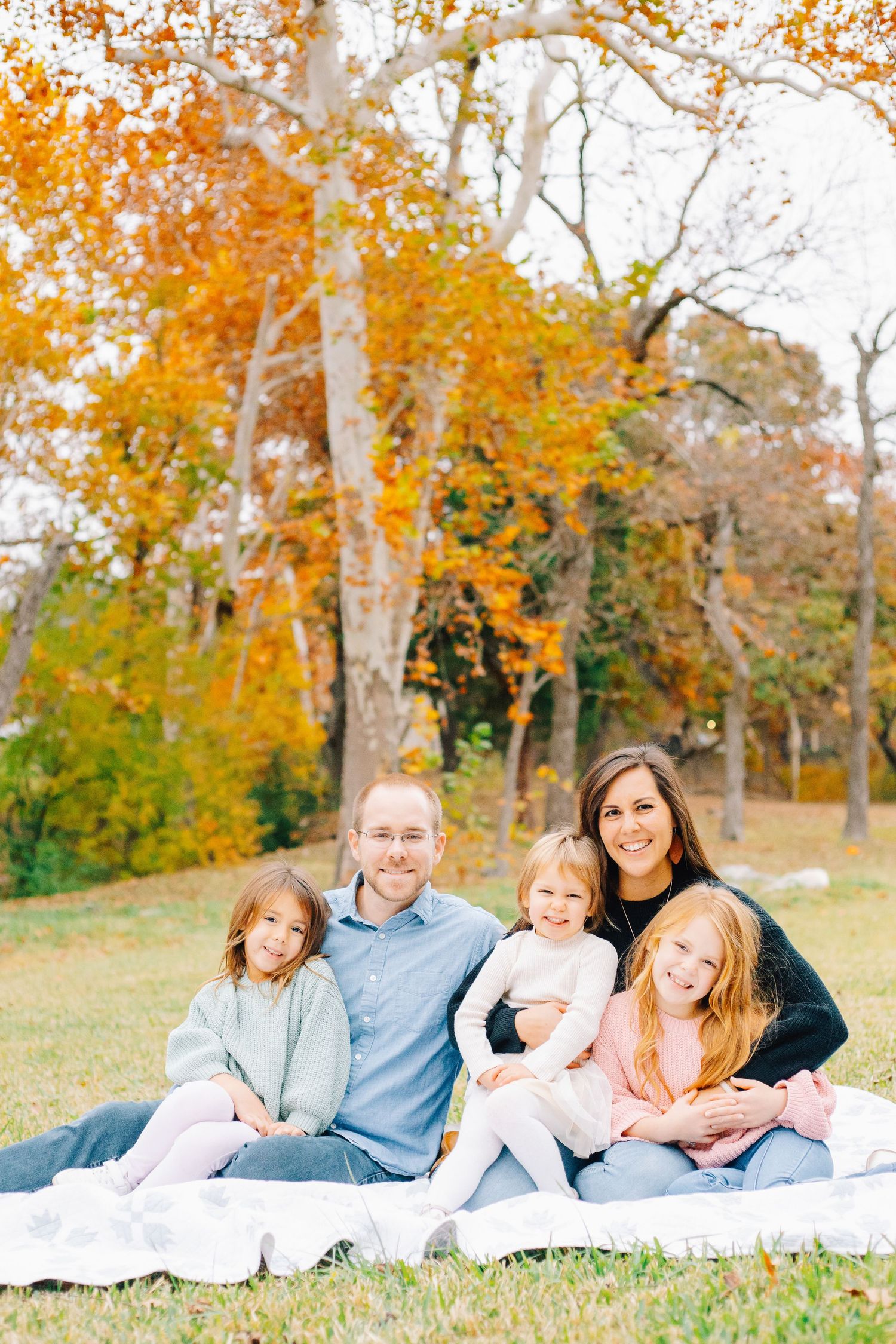 Family relaxes together on white blanket during autumn outdoor photoshoot.