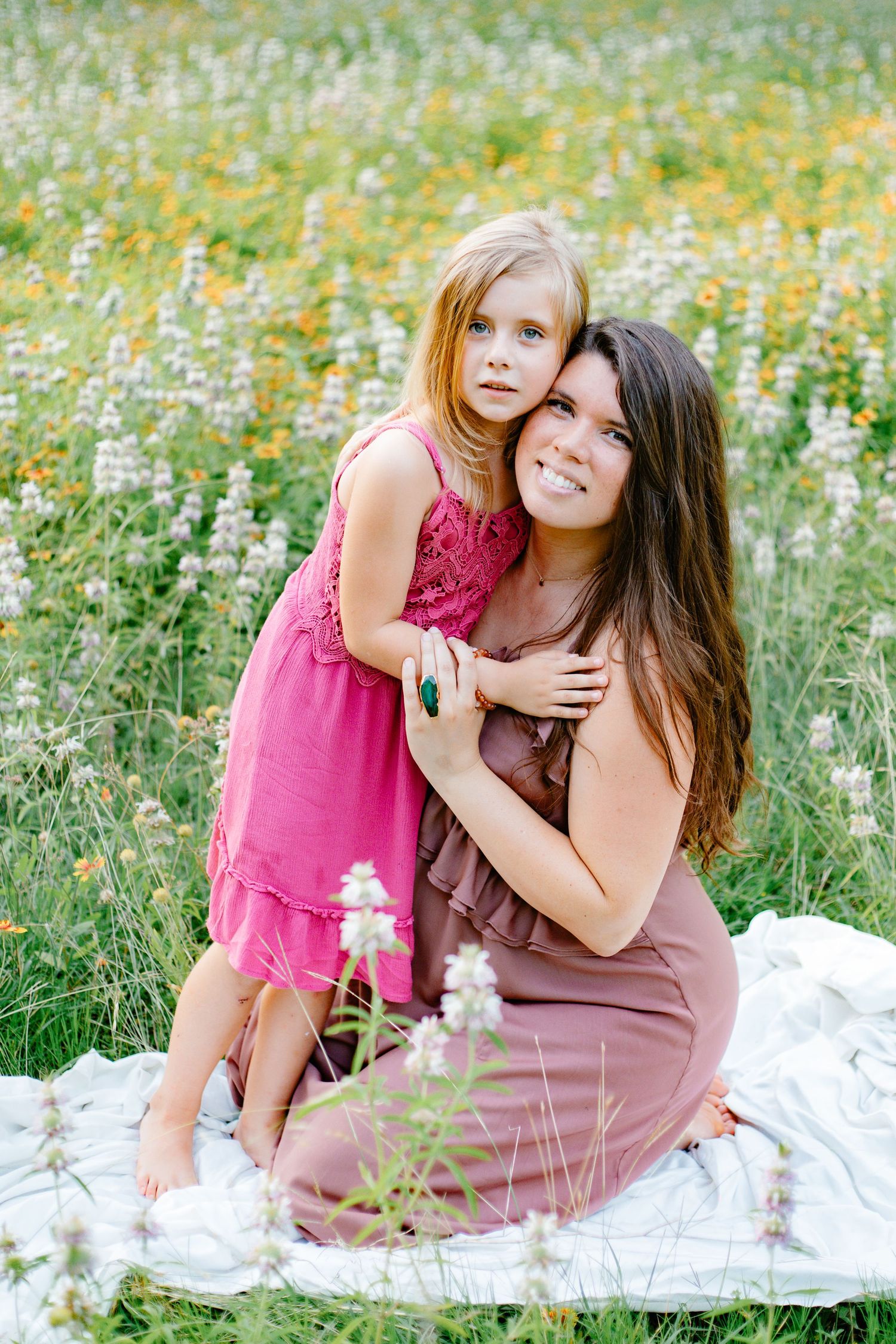 Smiling pregnant woman in mauve dress sits in wildflower field hugging blonde daughter in pink dress.