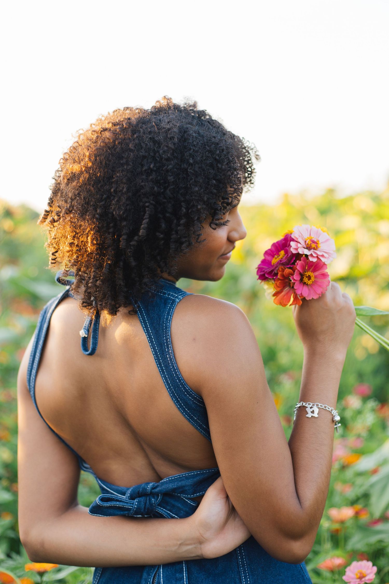 A woman with curly hair in a denim backless dress smells pink flowers in a colorful garden.
