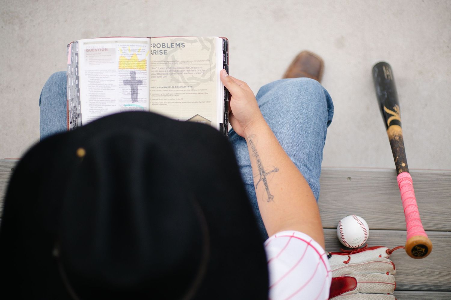 Person sitting on bench reading a book with a baseball bat, ball, and black hat nearby.