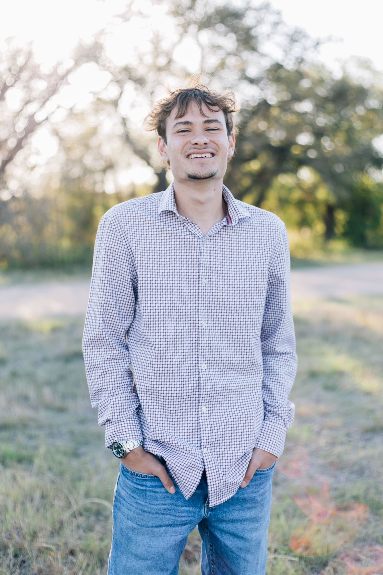 Smiling young man in patterned button-up shirt and jeans standing outdoors in a field with trees in background. Senior Portraits TX