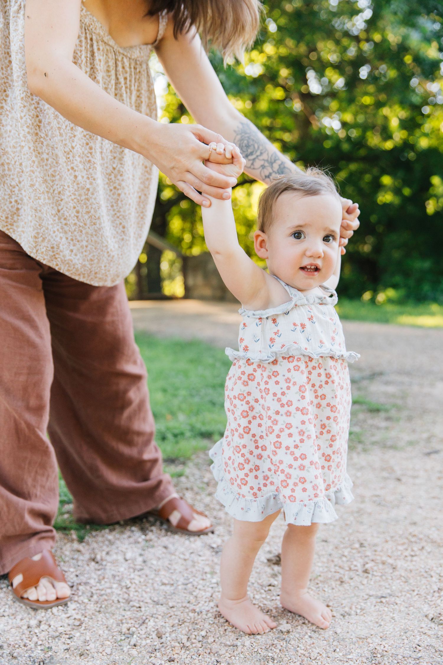 A mother helps her barefoot toddler girl in a floral dress take first steps outdoors in a sunny park.