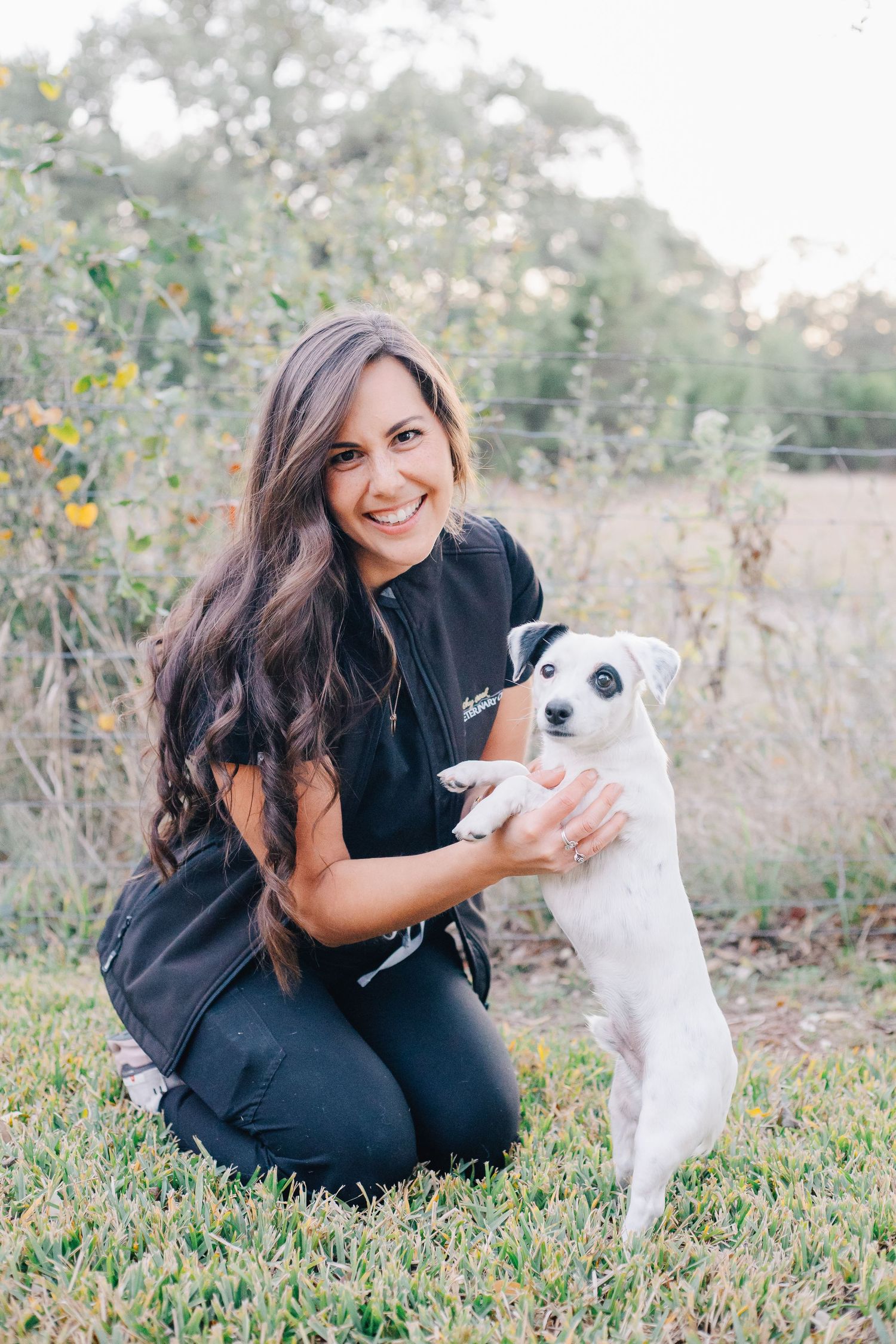 A woman in black plays with her small white dog on green grass.