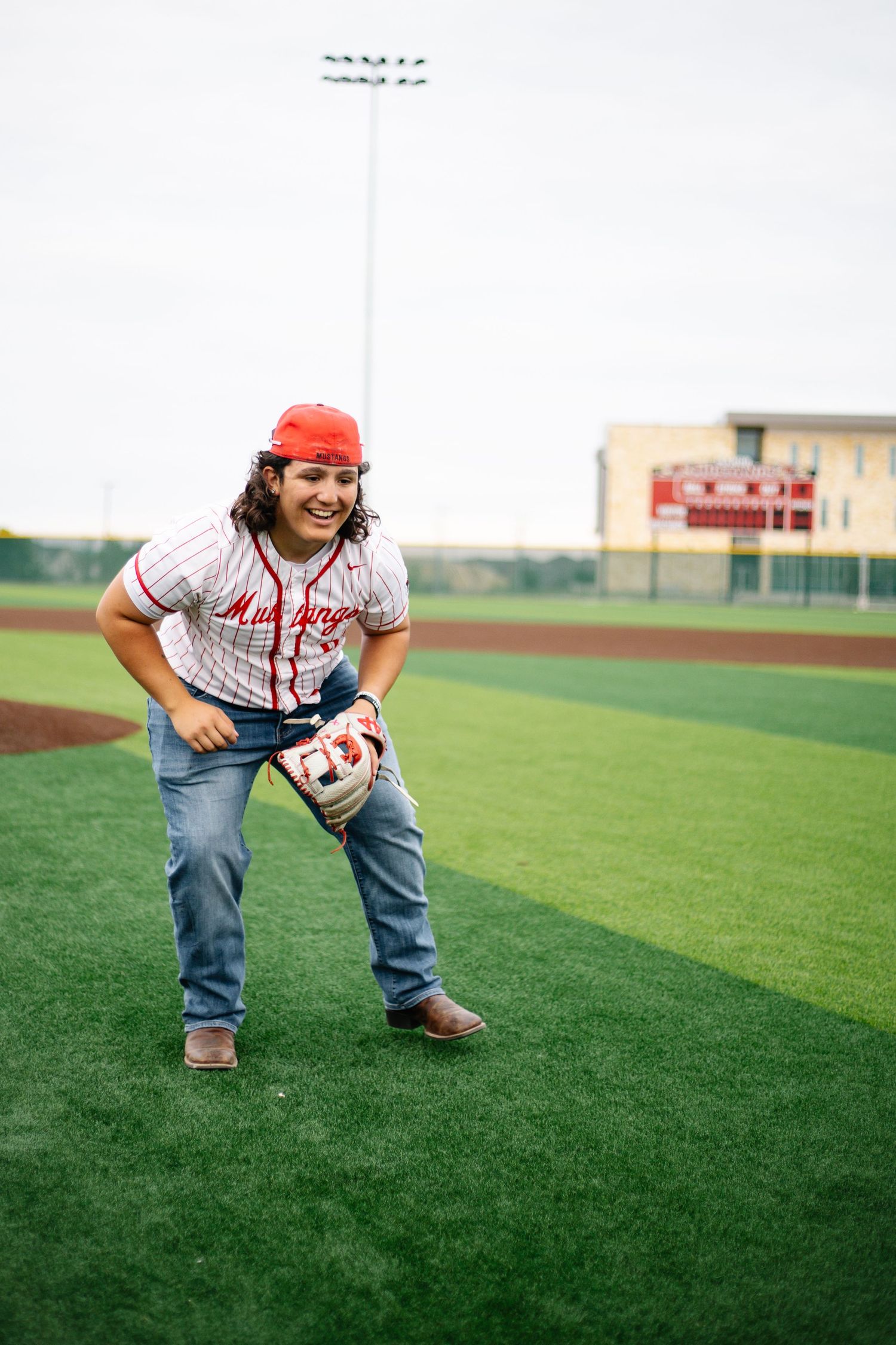 Young woman in baseball uniform and red cap smiling on green turf field holding a glove. Senior Portraits Austin TX
