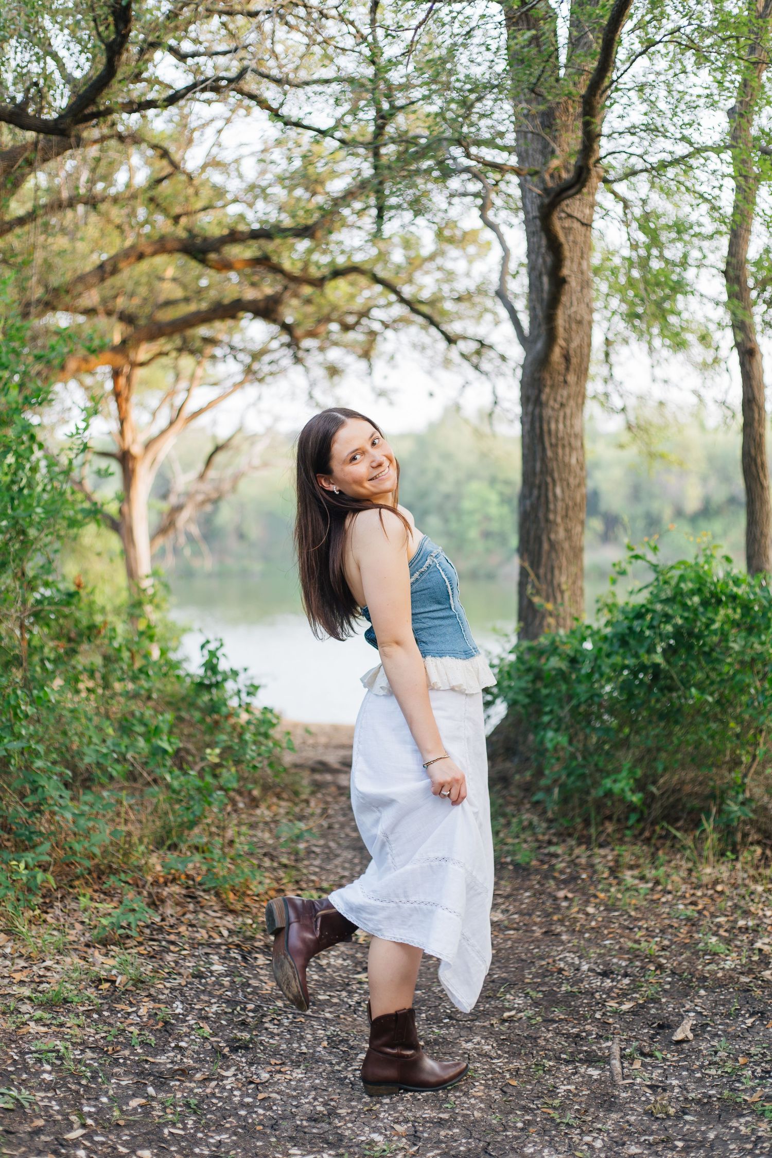 Young woman in denim top and white skirt with brown boots walking on woodland path by lake. Senior Portraits Round Rock TX