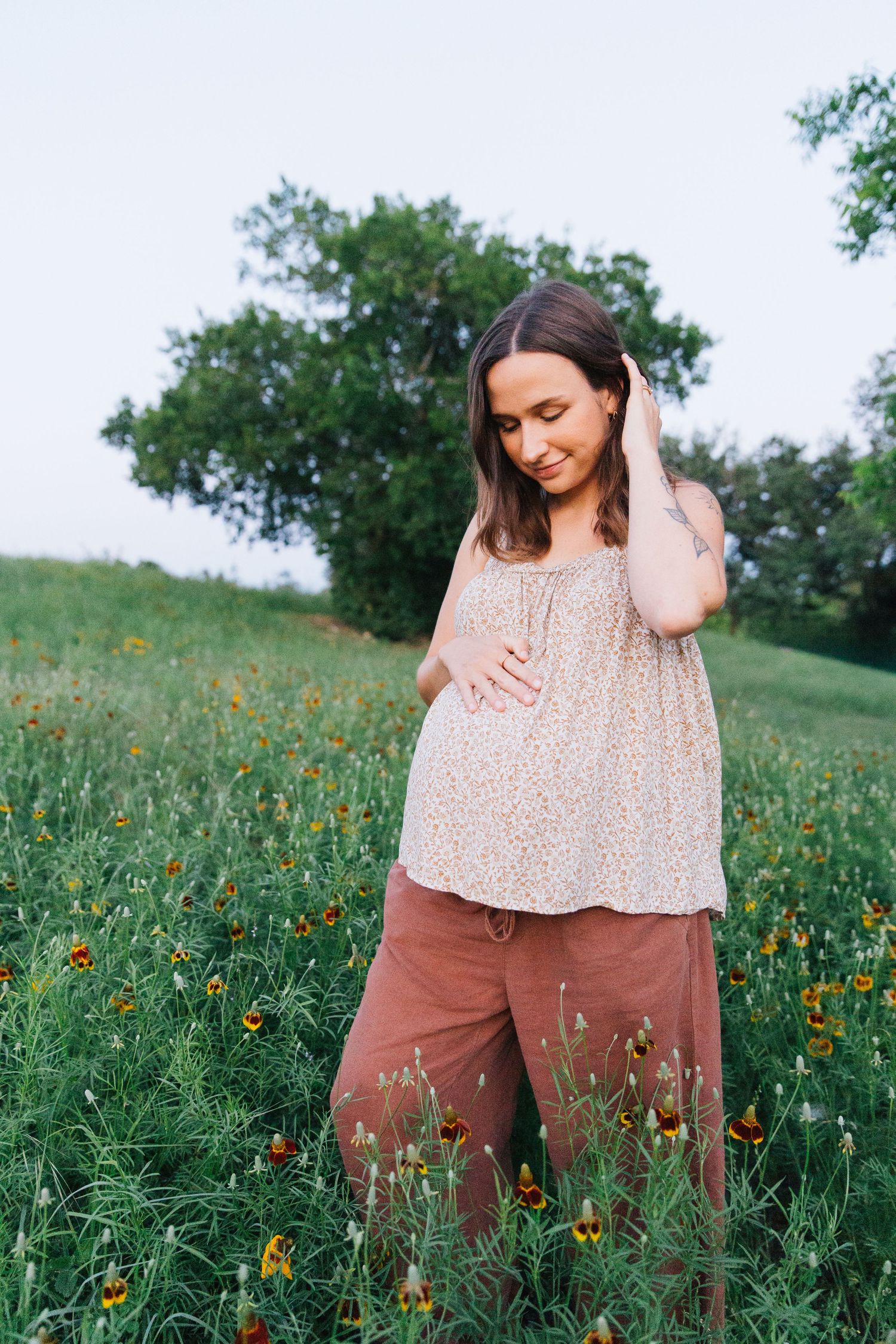 Pregnant woman in floral top and rust pants standing in a wildflower field, gently cradling her baby bump outdoors.