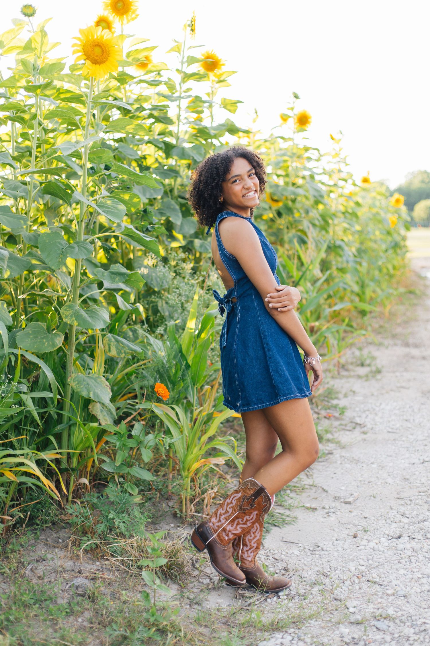 Smiling young woman in denim dress and cowboy boots posing beside tall sunflowers on a sunny dirt path.