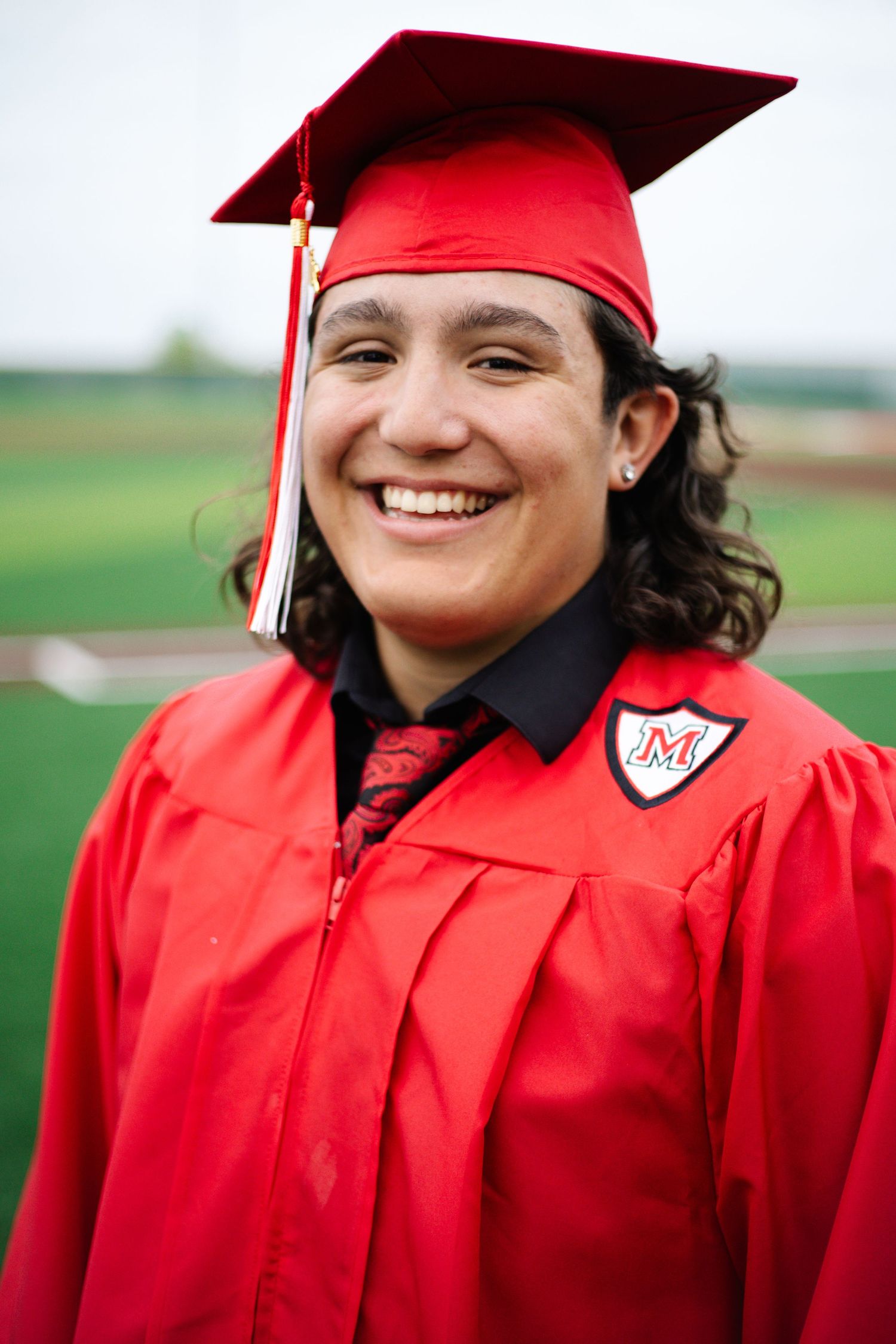 Smiling graduate wearing red cap and gown with M emblem poses outdoors on green field.