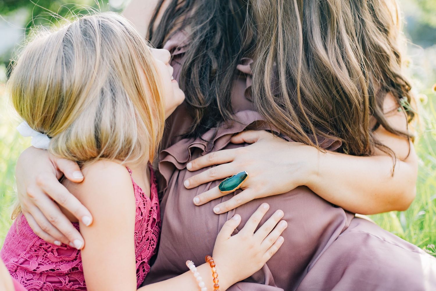 Young blonde girl in pink dress hugging pregnant mother during outdoor family maternity photo session.