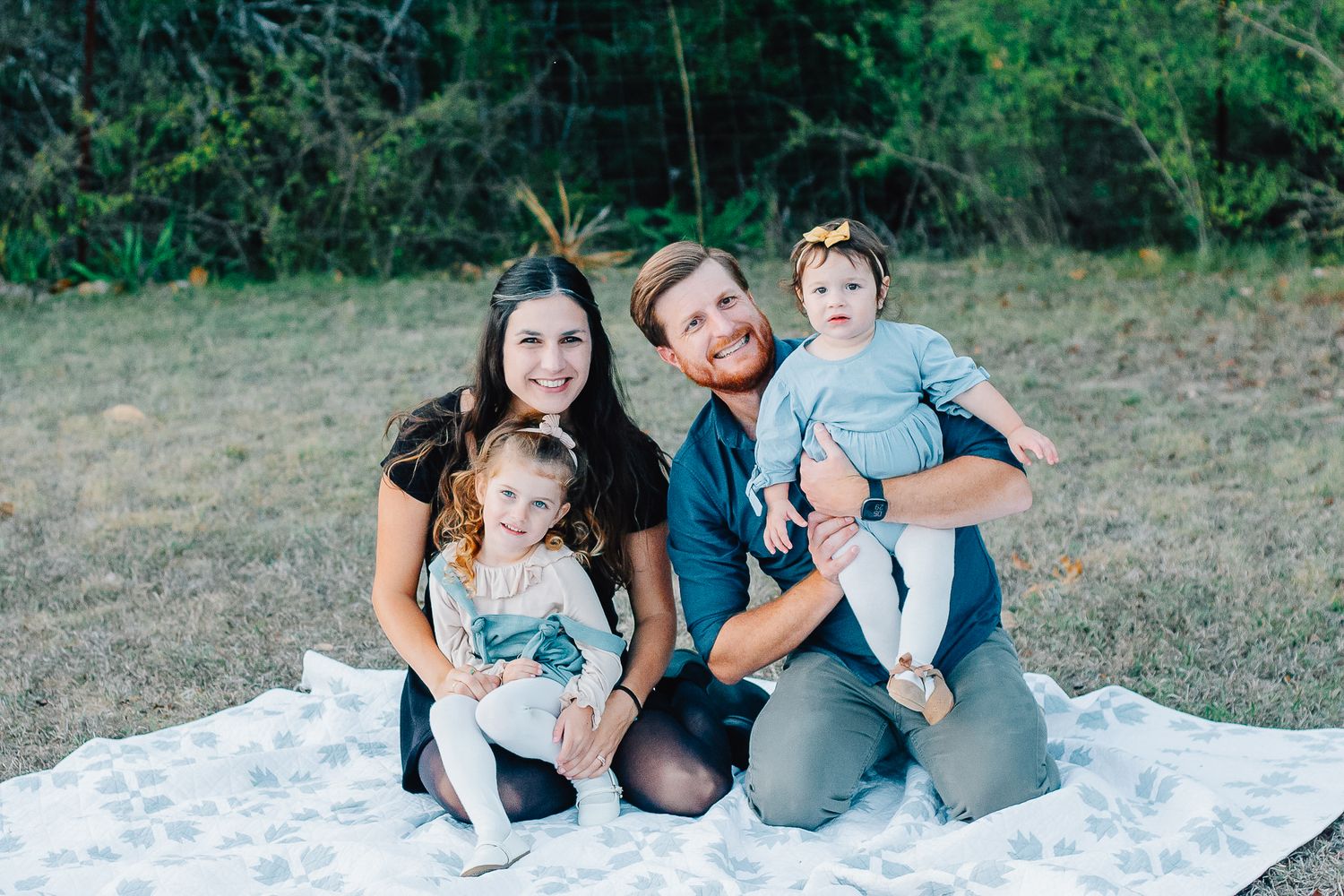Family group sitting together on white blanket outdoors during photo session.