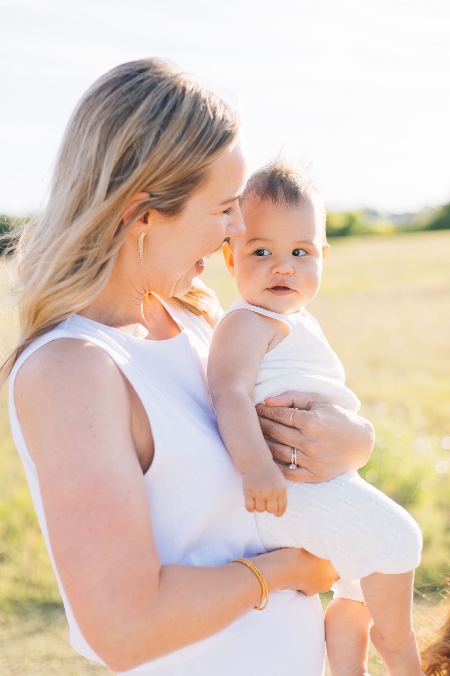 A blonde woman in white dress holds a cute baby outdoors in a sunny field.