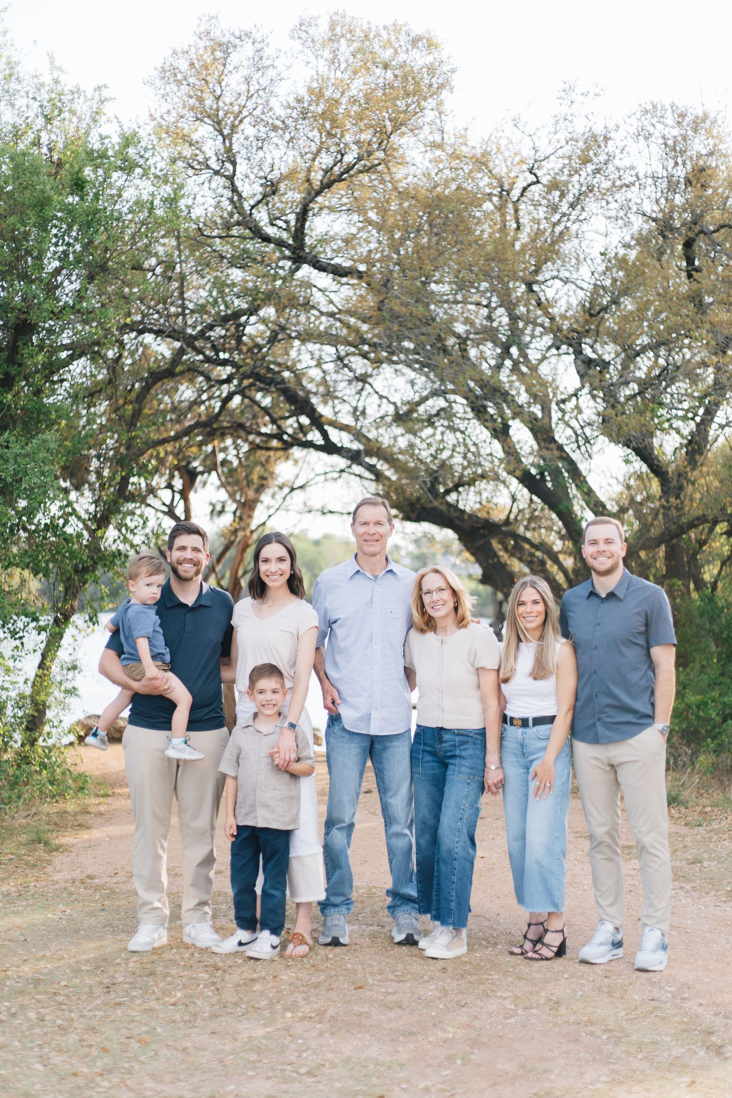 Large family group portrait outdoors near a lake surrounded by oak trees on a sunny day.