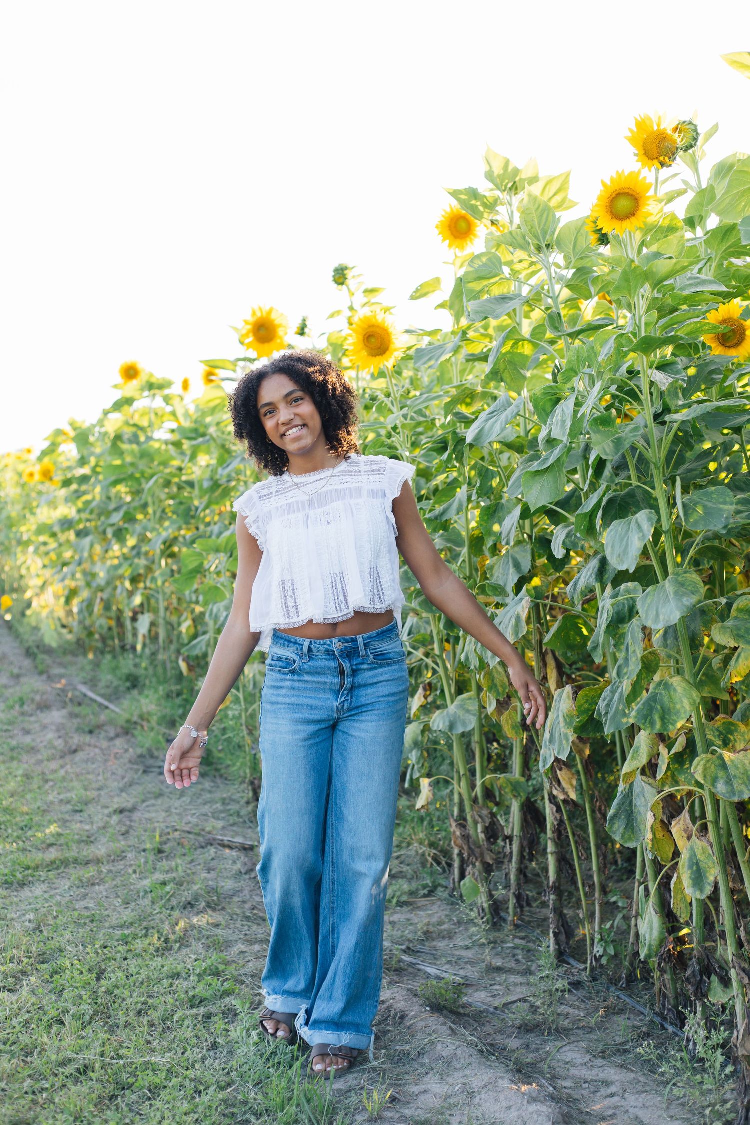 Young Black woman in white top and flared jeans walking along sunflower field path, smiling outdoors.