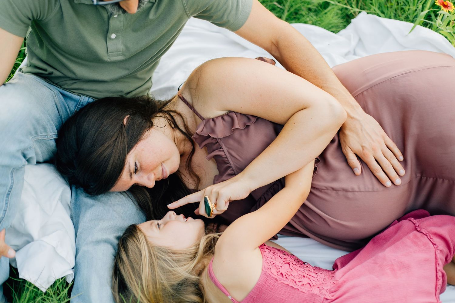 Two women and a man relaxing on grass, with a pregnant woman in a mauve dress being embraced outdoors.