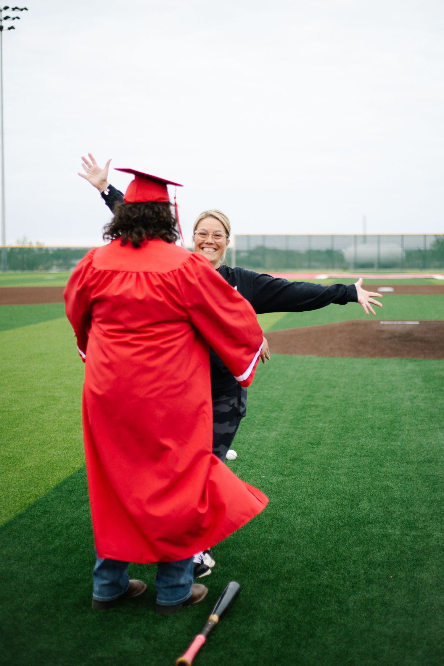 Graduate in red cap and gown celebrates on baseball field with smiling woman in joyful outdoor ceremony.