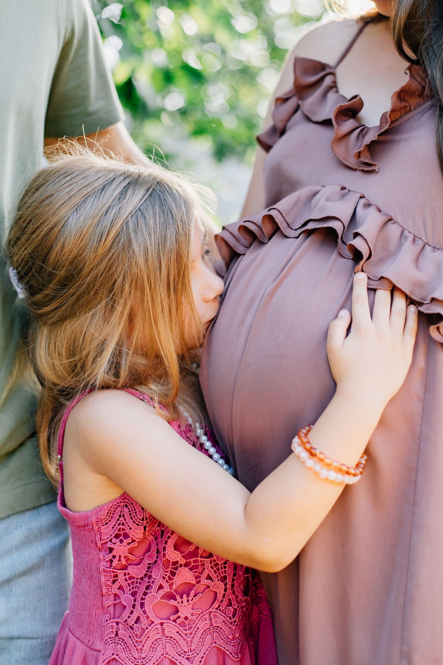 Little girl in pink dress kissing pregnant mothers baby bump outdoors during family maternity photography session.