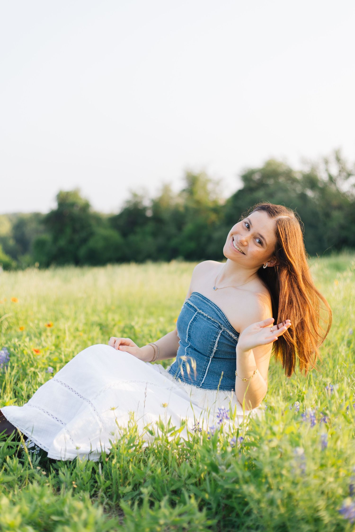 Beautiful woman with long red hair in blue denim top and white skirt relaxing in a sunny wildflower field. Senior Portraits Round Rock TX