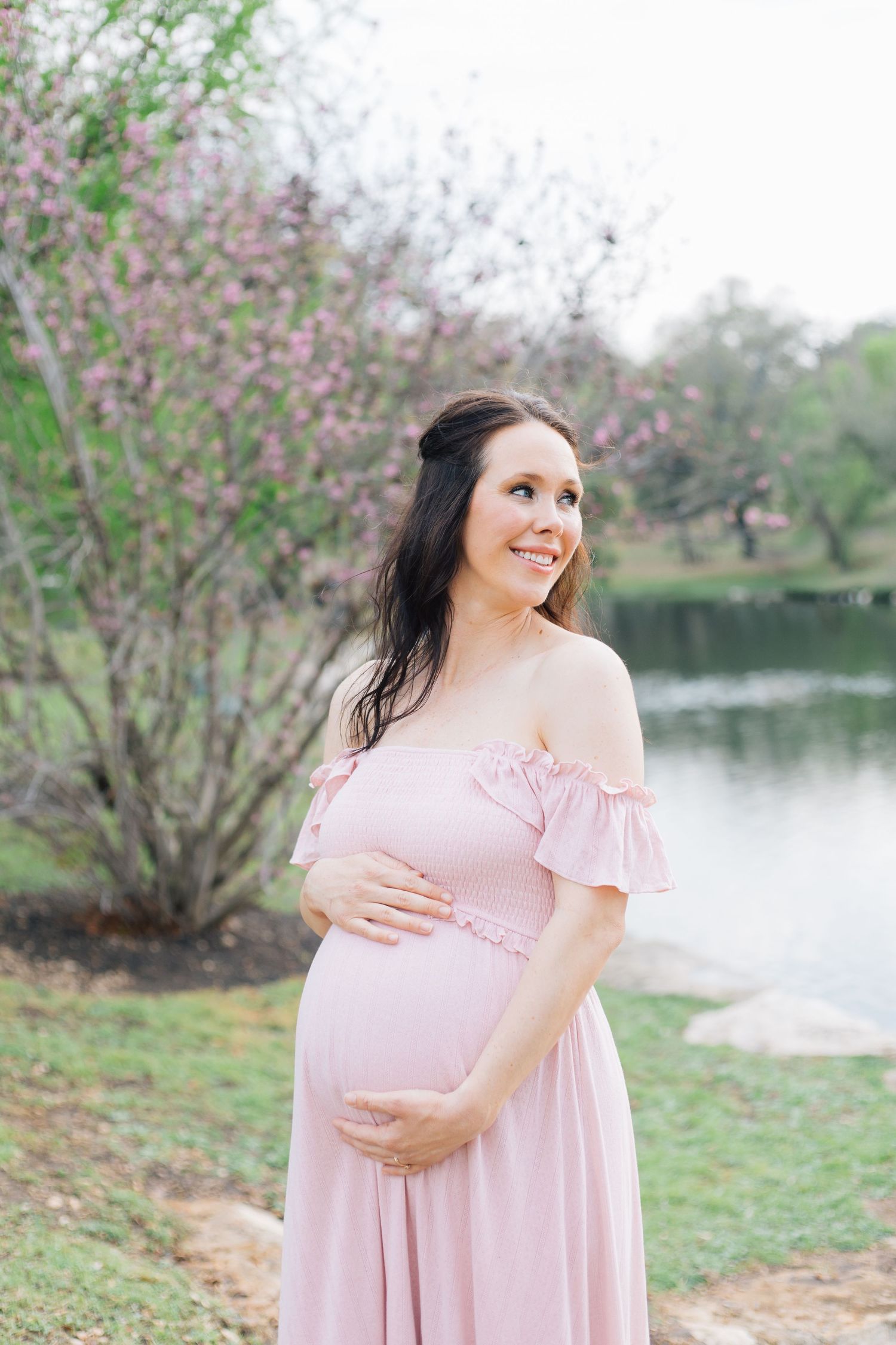 A woman in a pink dress cradles her baby bump while standing near blooming trees by a lake.