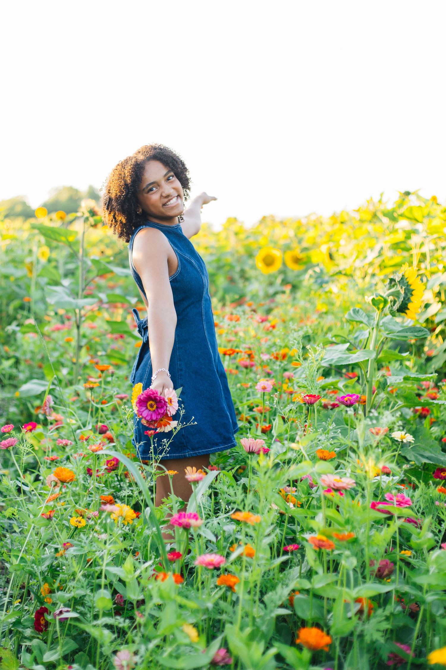 A smiling woman in a blue dress holds colorful flowers while standing in a vibrant field of zinnias and sunflowers.