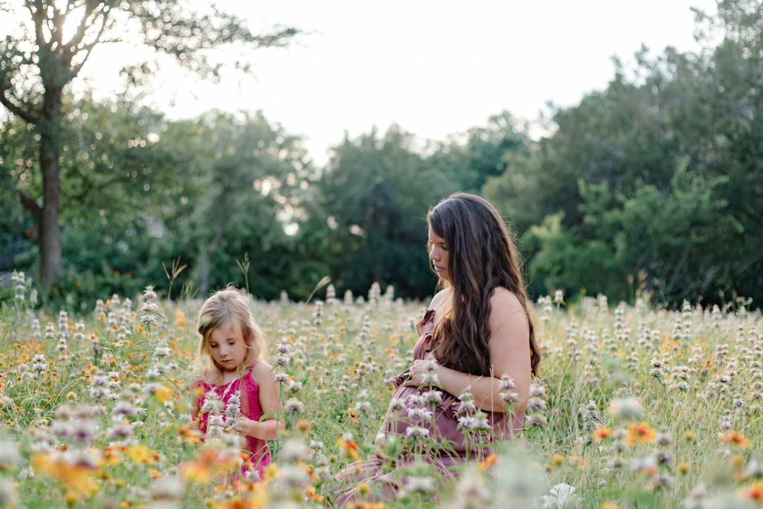 A mother and young daughter picking wildflowers together in a sunny meadow surrounded by trees.