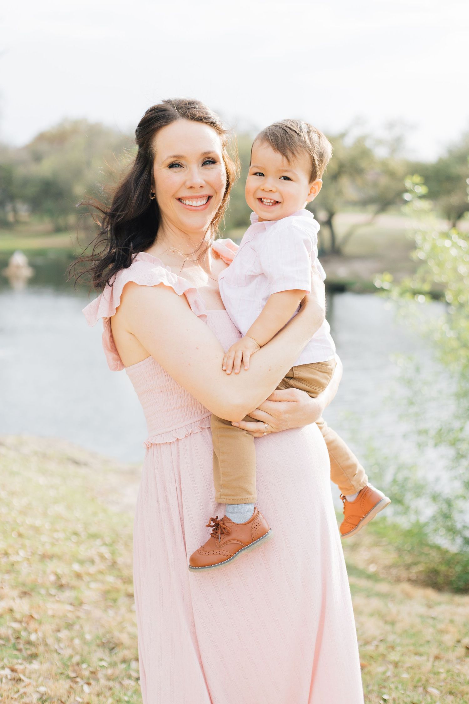 A woman in a pink dress holds a small child while standing near a waterfront during a spring day.