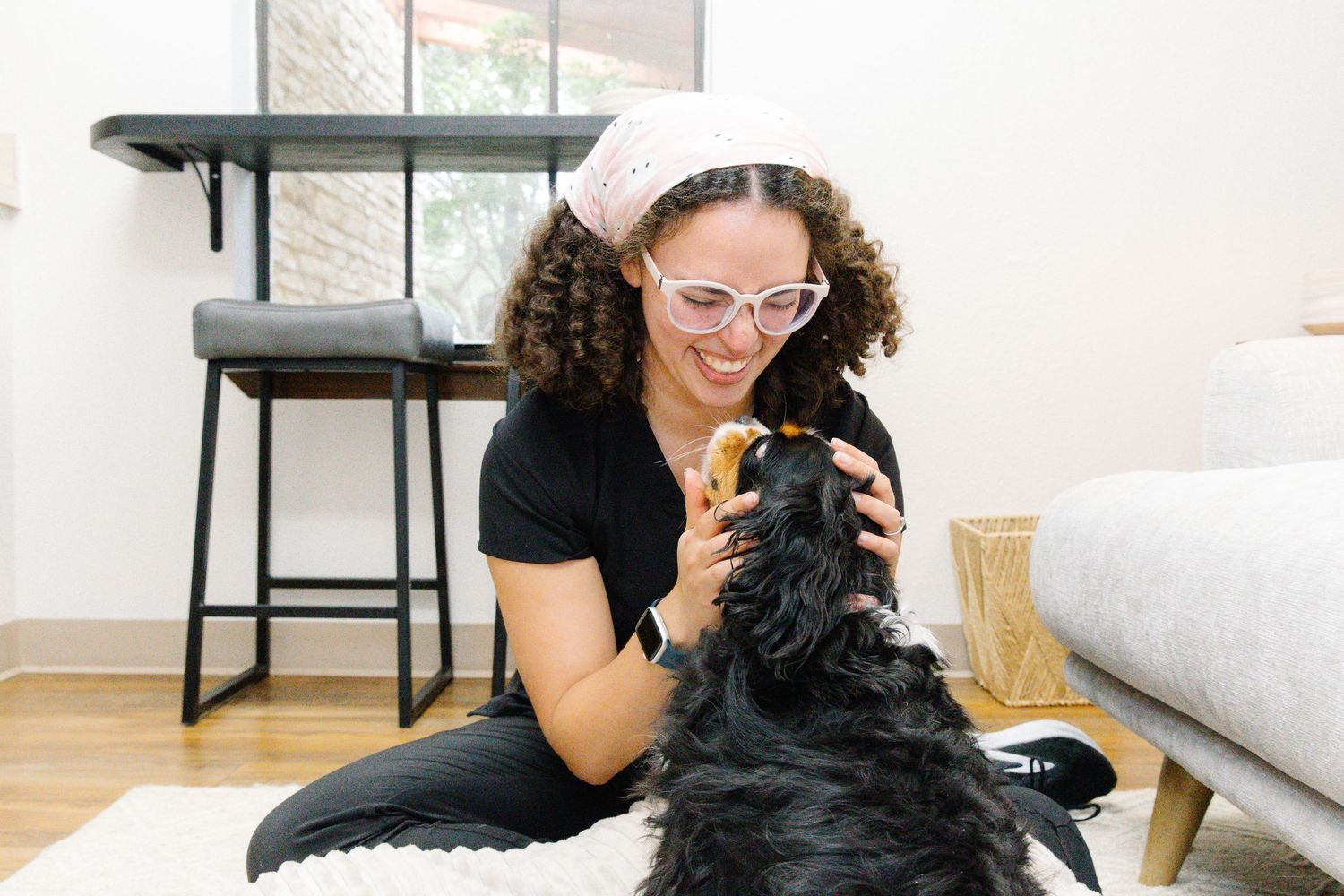 Woman with curly hair and pink glasses plays with black dog on floor in bright modern living room.