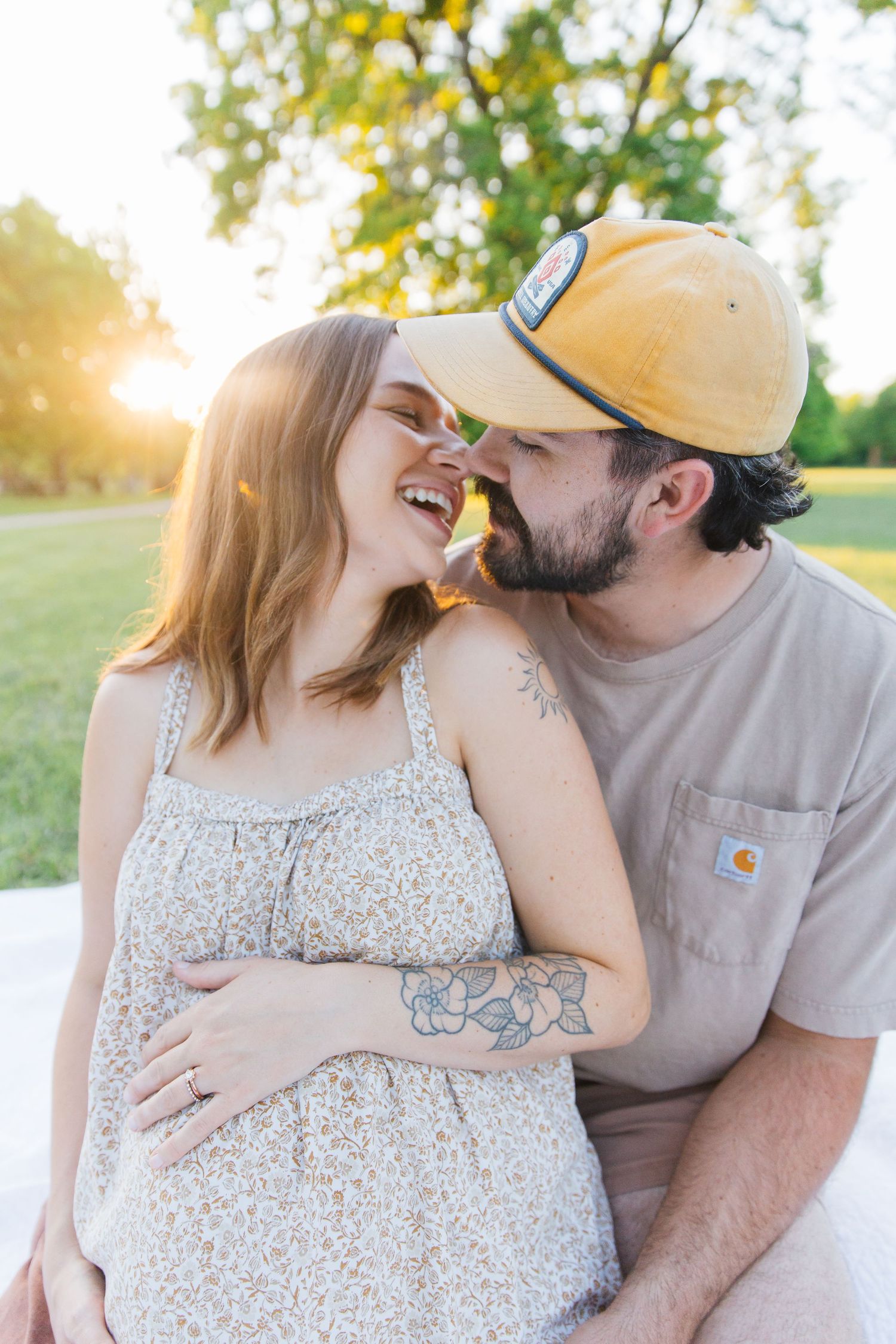 A bearded man in a yellow cap kisses a laughing pregnant woman outdoors at golden hour.