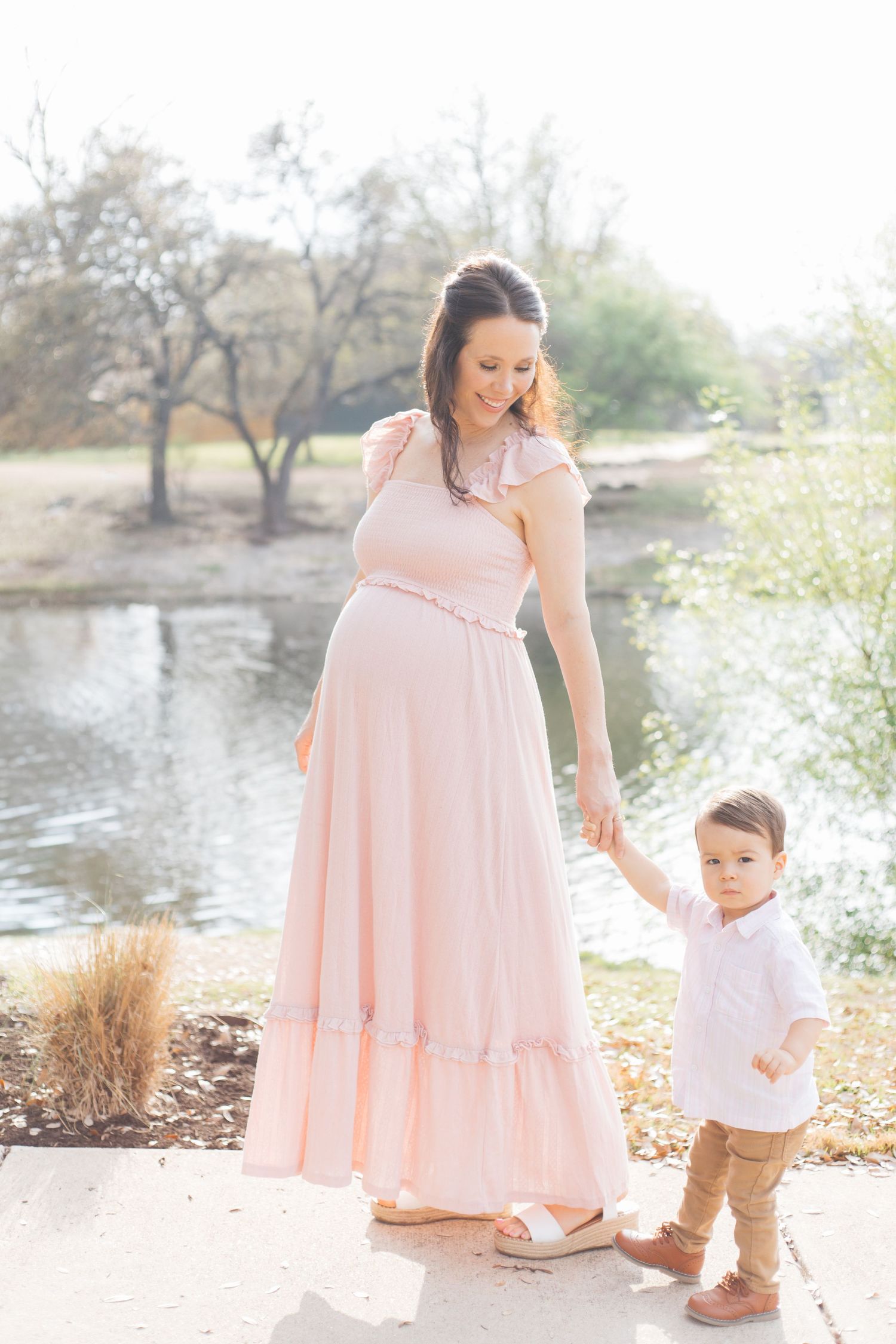 A woman in a flowing pink dress plays with a small child near a lake during a spring photoshoot.