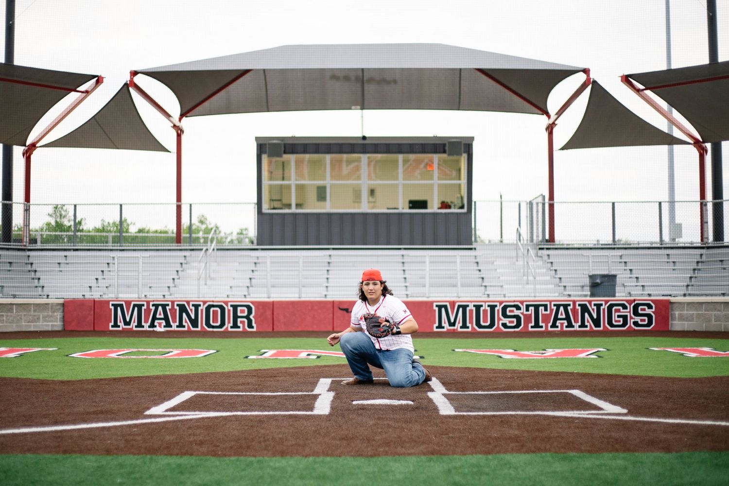 Student poses on baseball field at Manor Mustangs stadium, kneeling on home plate in front of red and white bleachers.